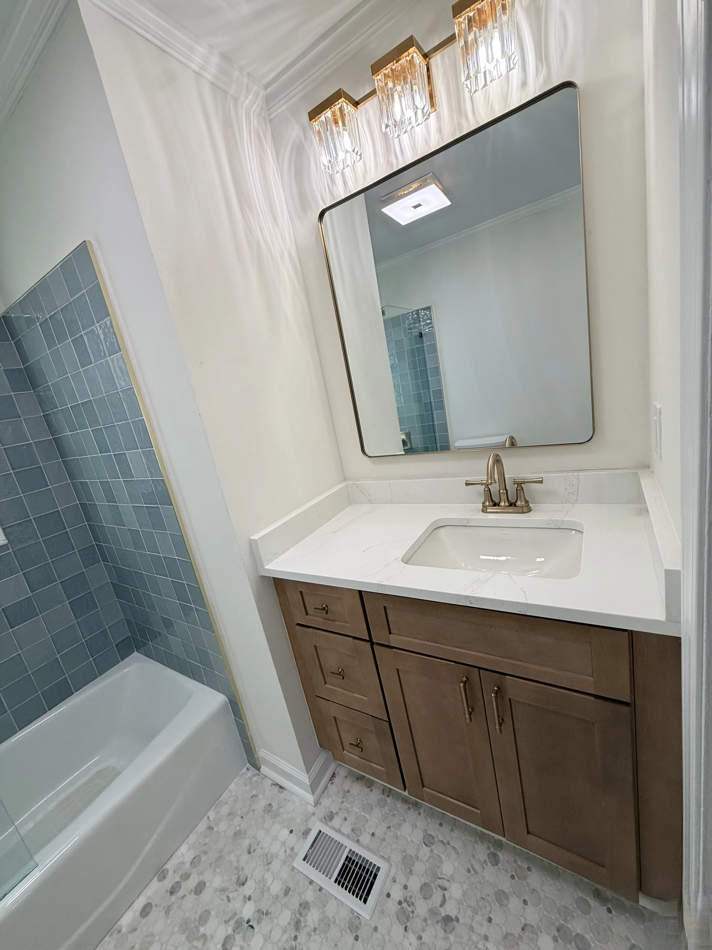 Bathroom with a wooden vanity, white marble countertop, rectangular sink, and gold faucet. Large mirror above the sink, modern light fixture with multiple crystal-style lights, and a blue-tiled shower with glass door on the left. White walls, ceiling with crown molding, and a floor vent.