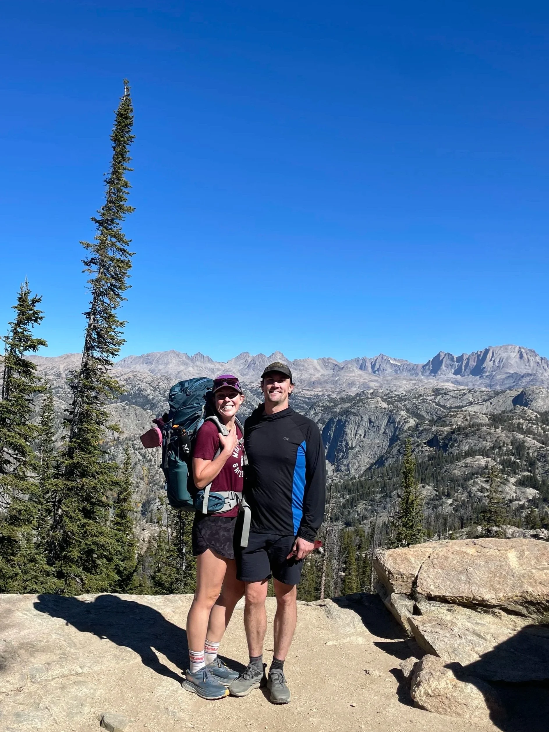 A smiling couple dressed in hiking gear, standing on a rocky trail with a scenic mountain landscape in the background, including tall trees, distant mountain peaks, and a clear blue sky.