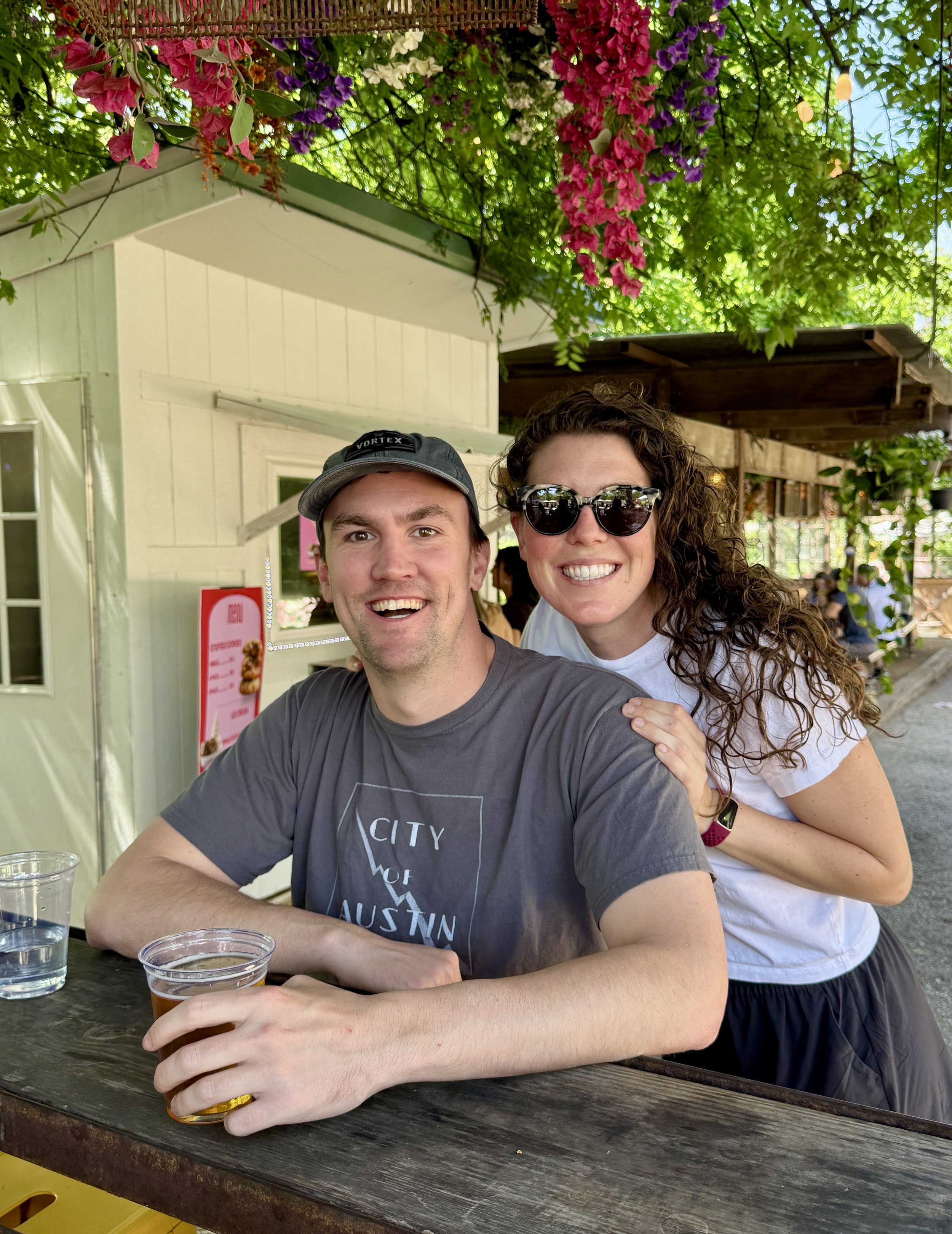 Two people, a man and a woman, are smiling at a camera while sitting at a wooden table outdoors under a lush green canopy with hanging pink, purple, and white flowers. The man is wearing a gray T-shirt with the words "City of Austin" and a dark cap, and the woman is wearing sunglasses and a white T-shirt. They appear happy and enjoying a casual outdoor gathering.