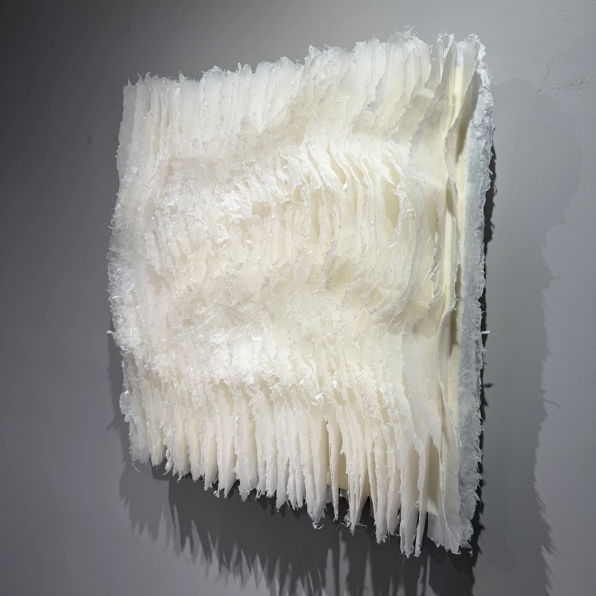 Textured white abstract art piece with layered, jagged, and wavy surface mounted on a gray wall.