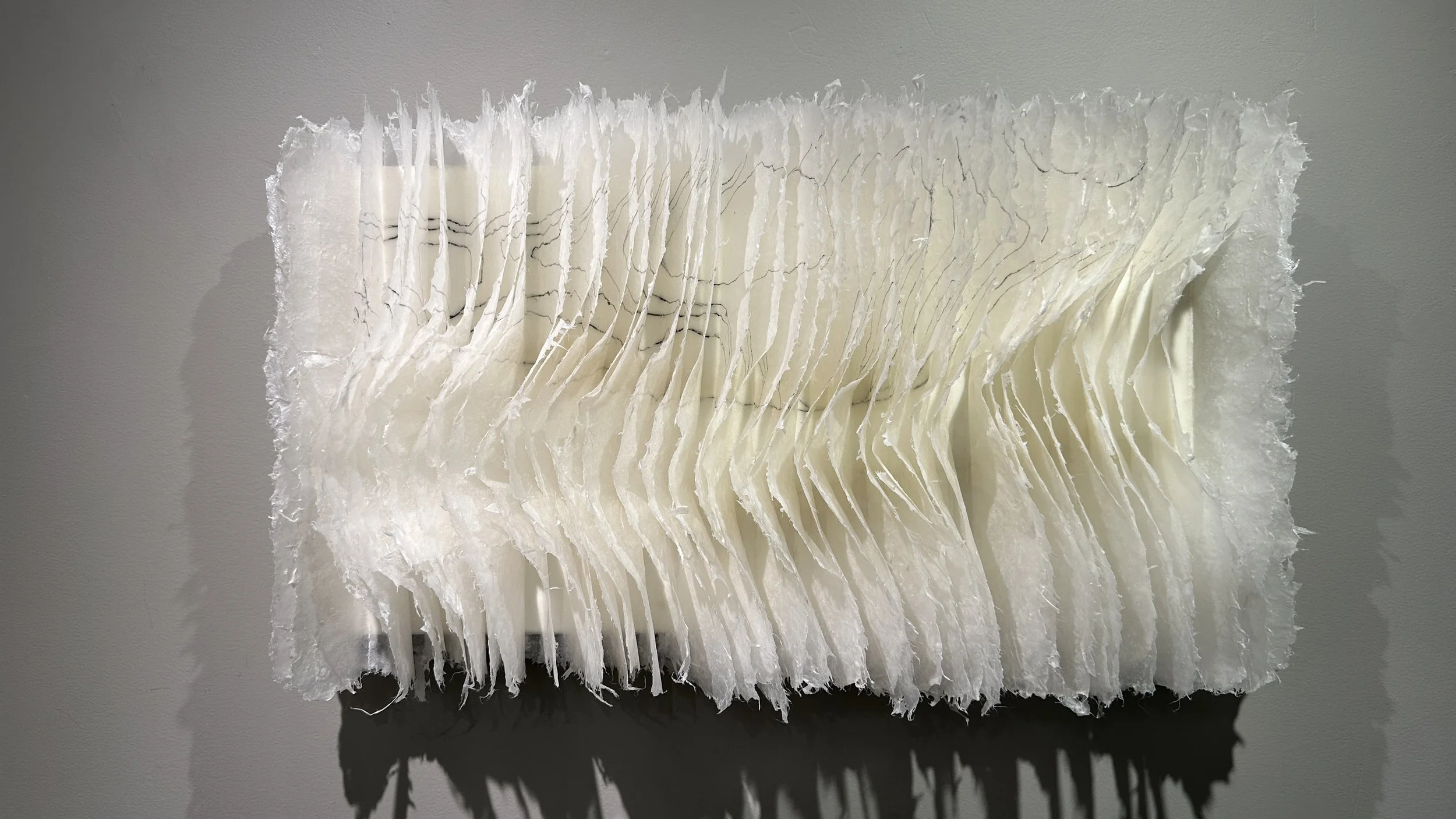 A white 3D textured wall sculpture with layered, irregular, vertically oriented paper-like strips casting shadows.
