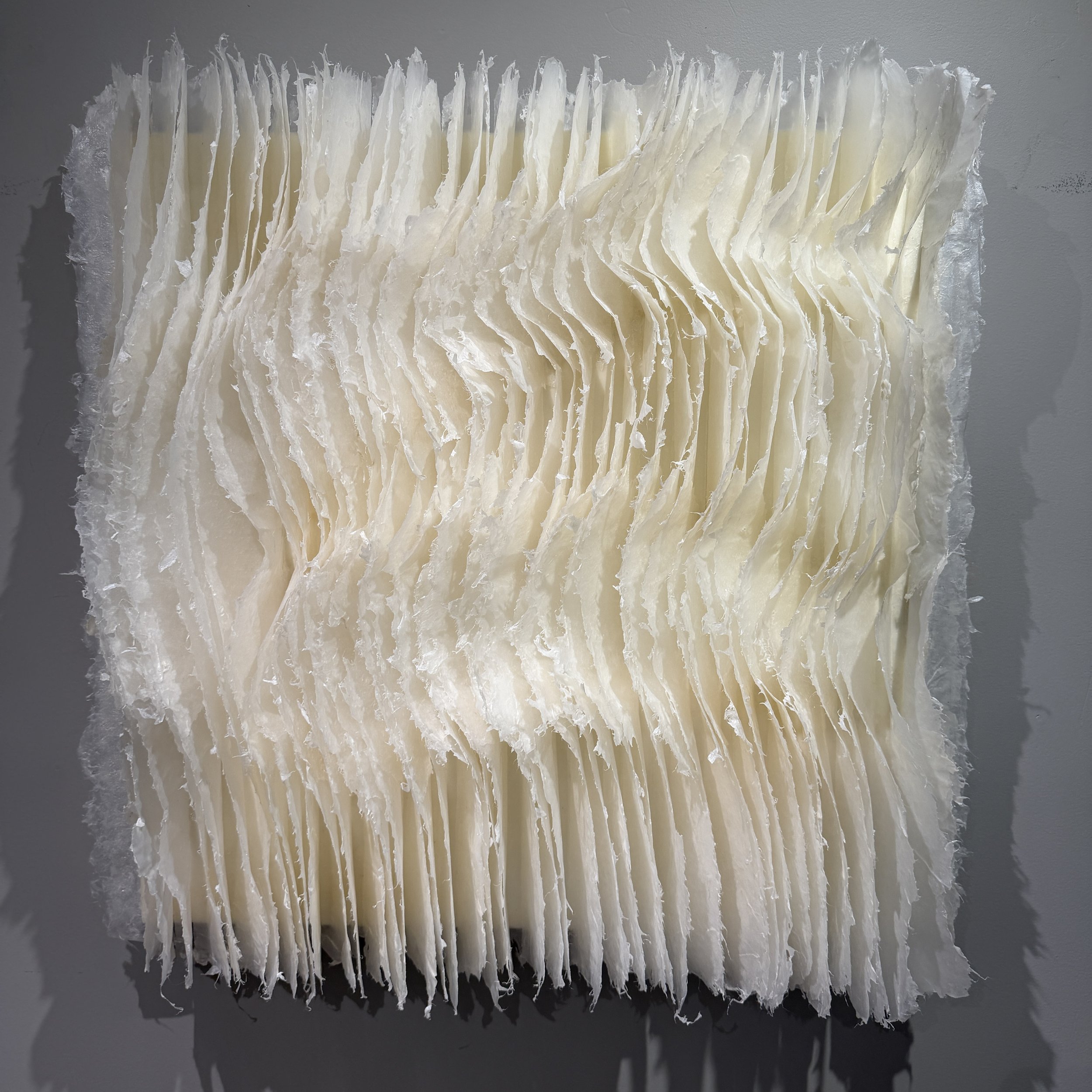 Close-up of a textured artwork made from layered, thin, white kazo pulp paper,  arranged in wavy, vertical folds.