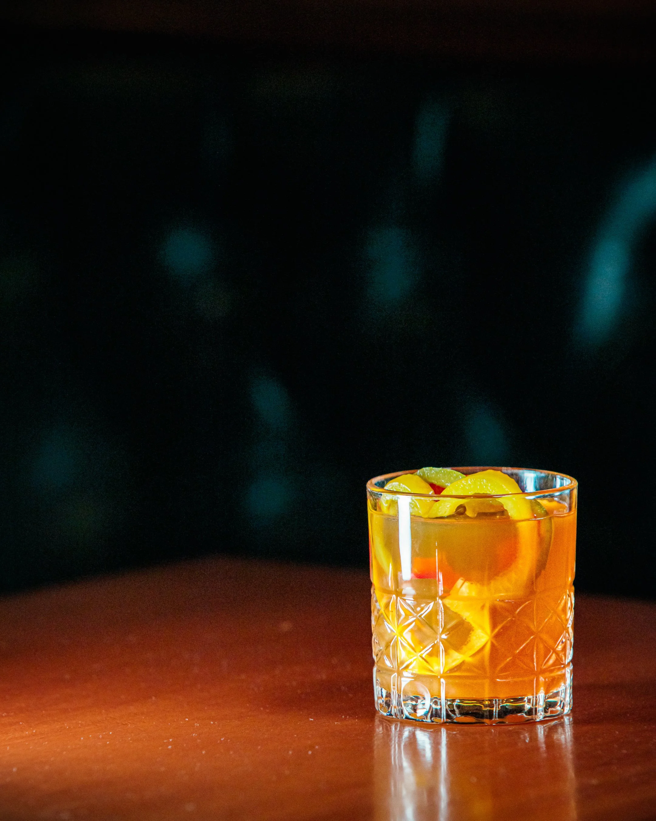 A close-up of an orange-colored cocktail with ice and fruit slices in a textured glass on a wooden surface with a dark, blurred background.