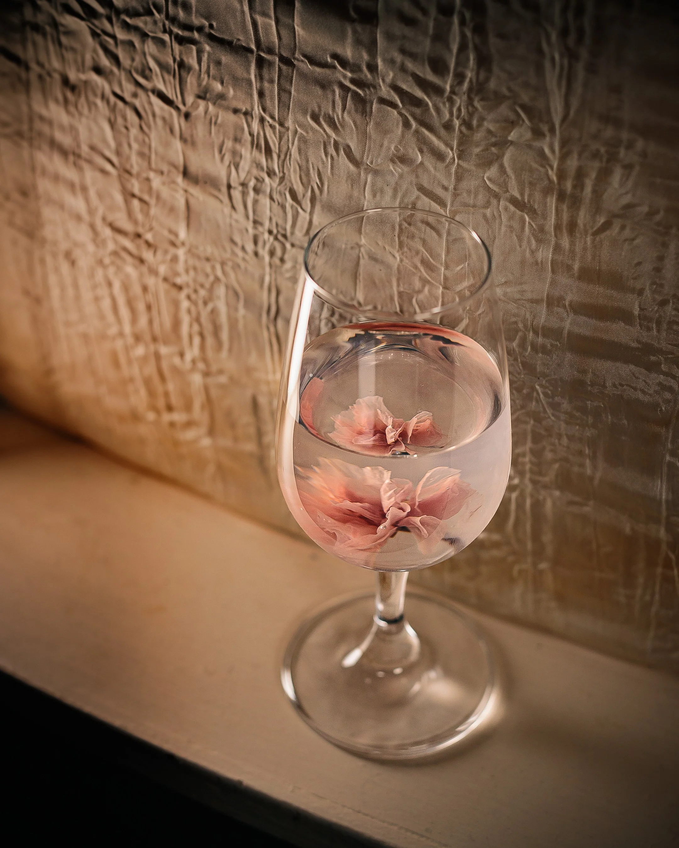 A wine glass filled with water and pink flower petals floating inside, placed on a white surface against a textured beige background.