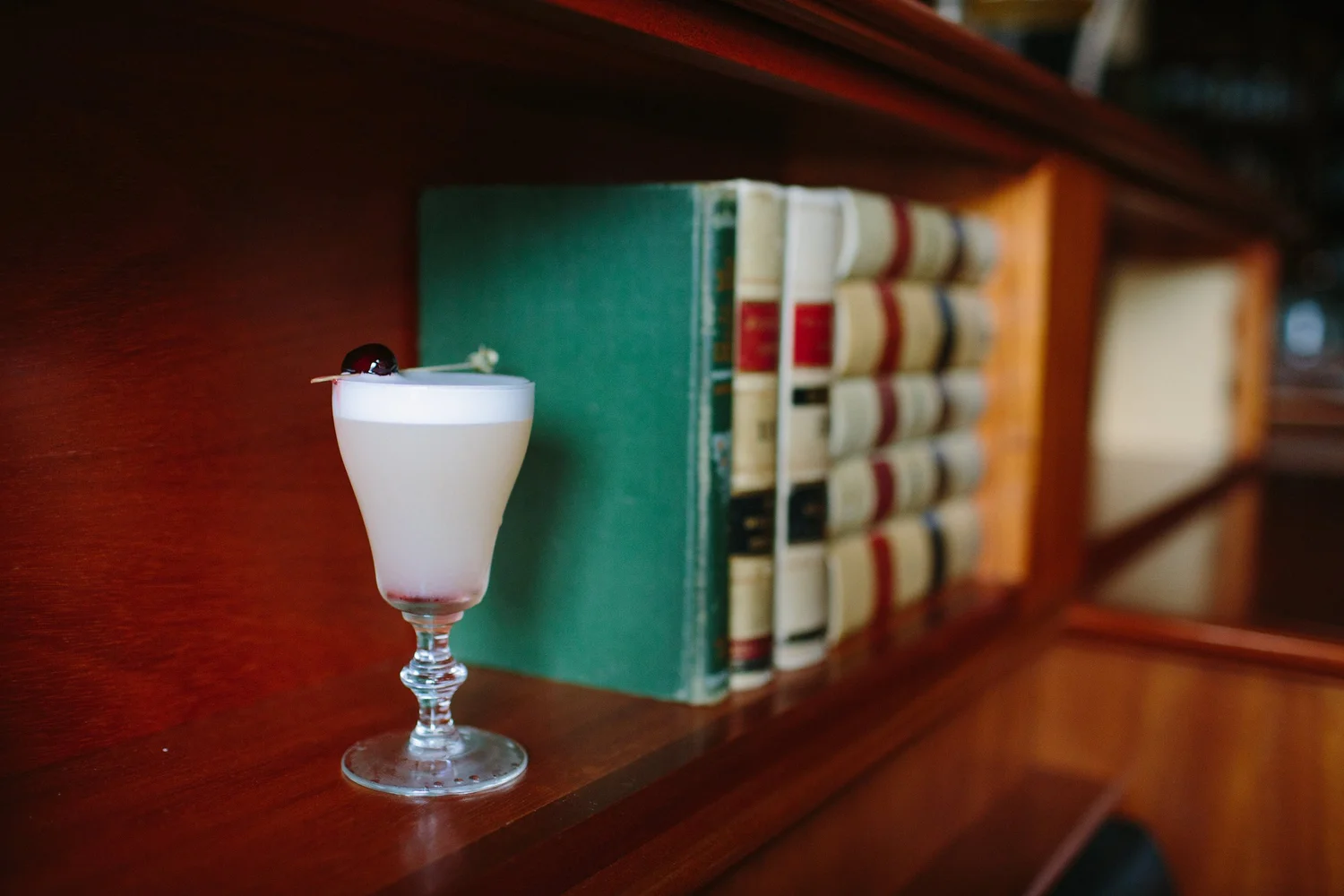 A frothy sour cocktail with a cherry garnish, served on the bar in front of law books at Tavern Law