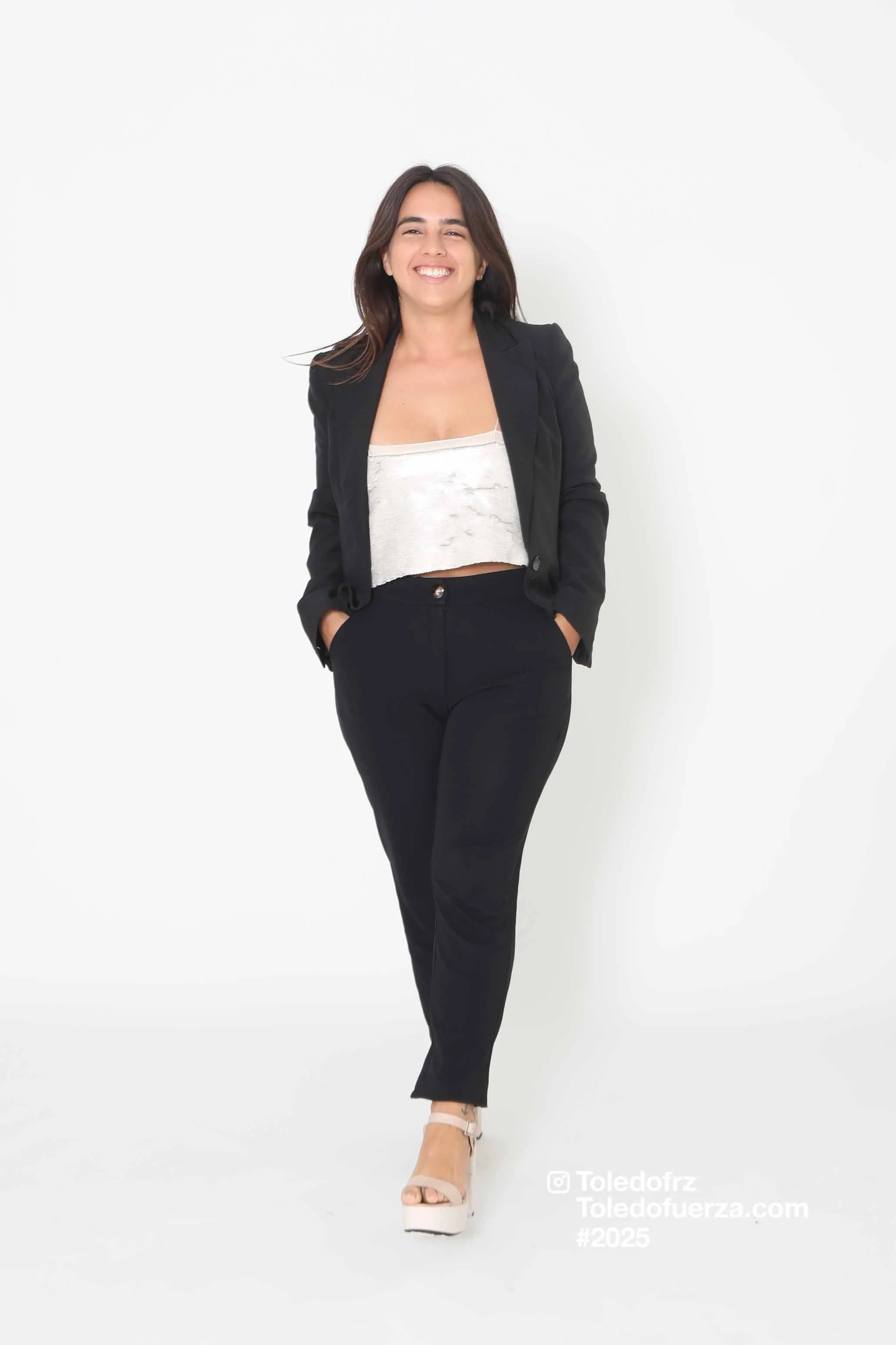Mujer sonriendo, vestida con blazer negro, top blanco, pantalones negros y sandalias beige, posando con las manos en los bolsillos, fondo blanco.
