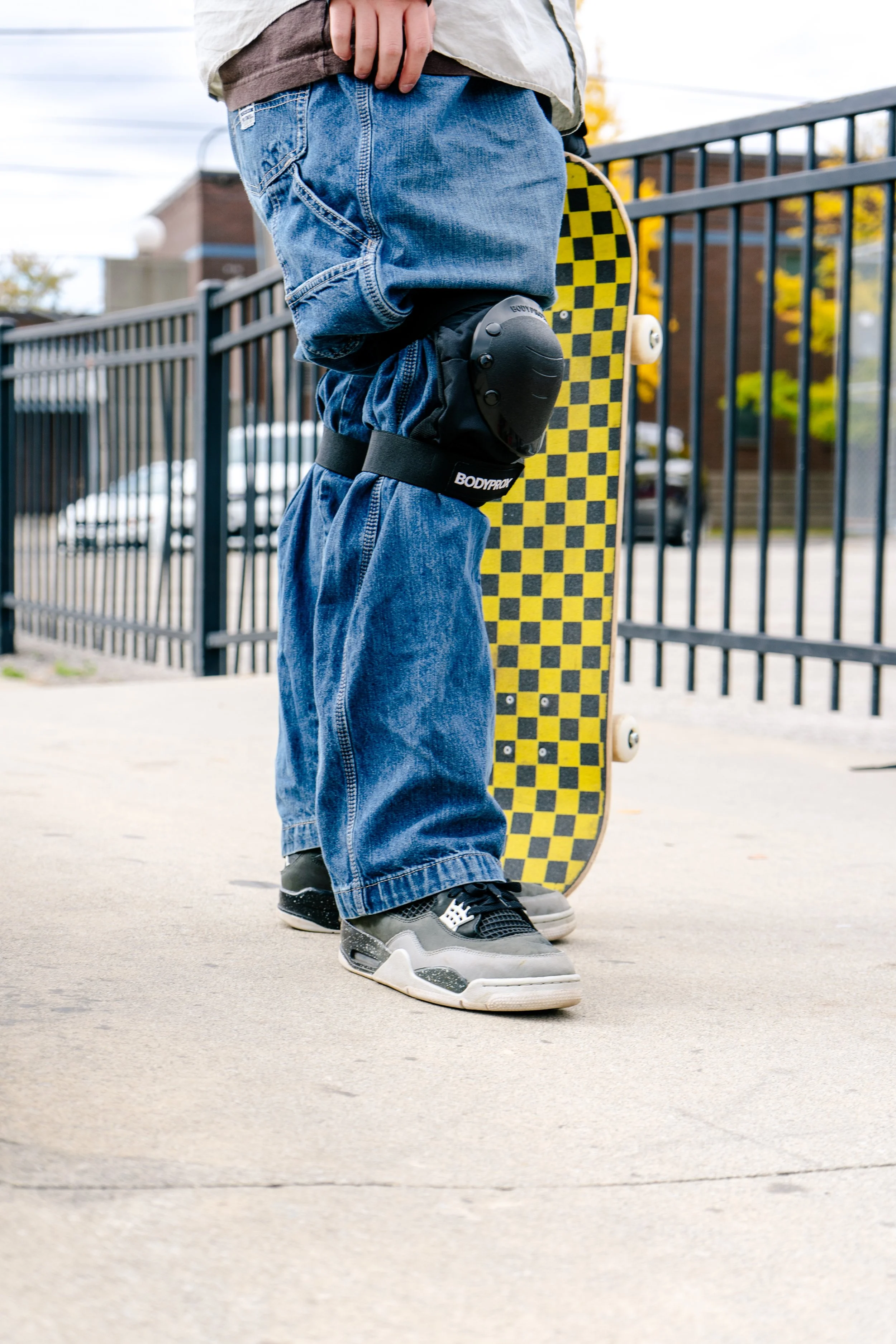 Close-up of a child standing on a sidewalk with a skateboard, wearing jeans, sneakers, and knee pads.