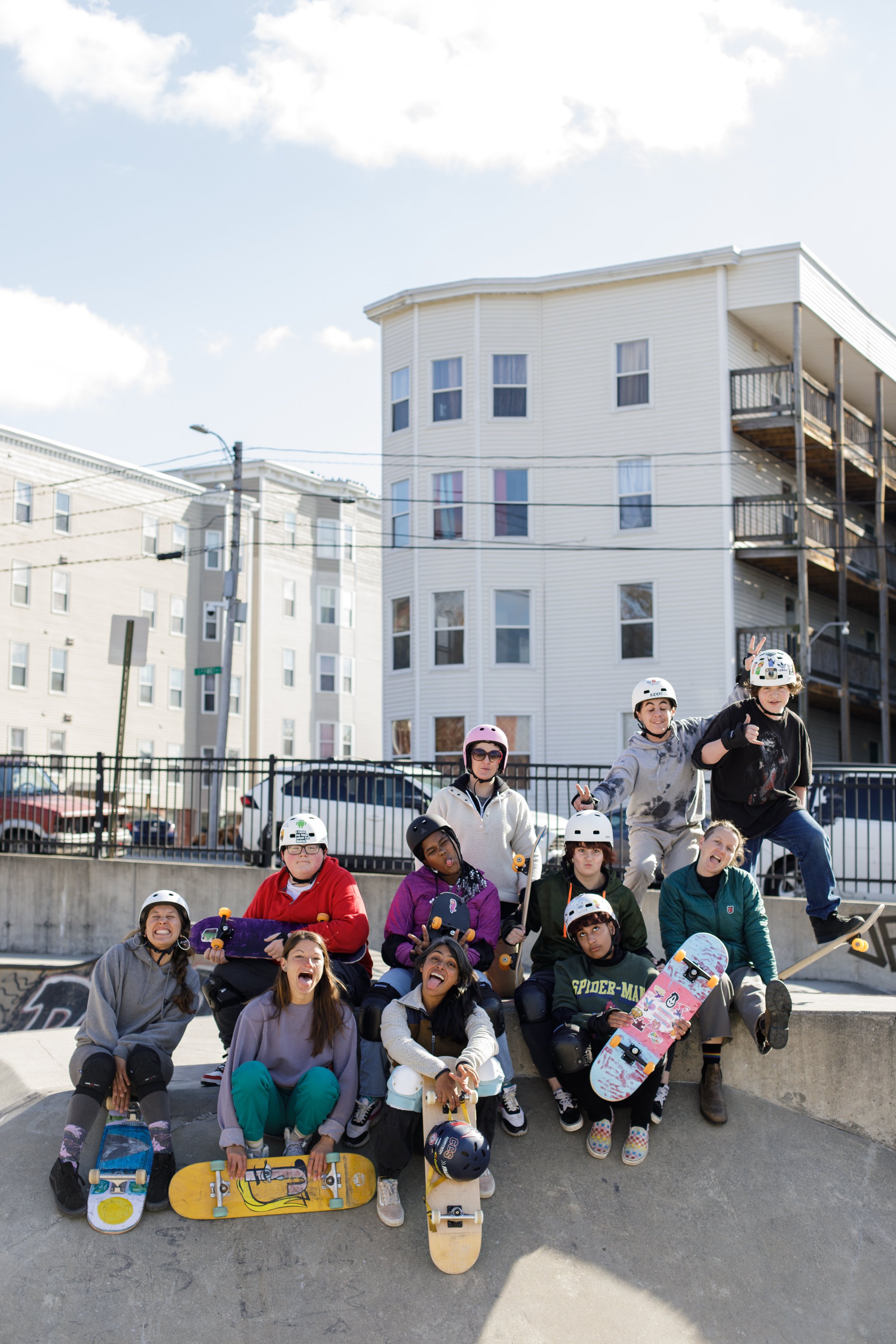 A group of young people at a skate park, some holding skateboards, wearing helmets, and making playful gestures.