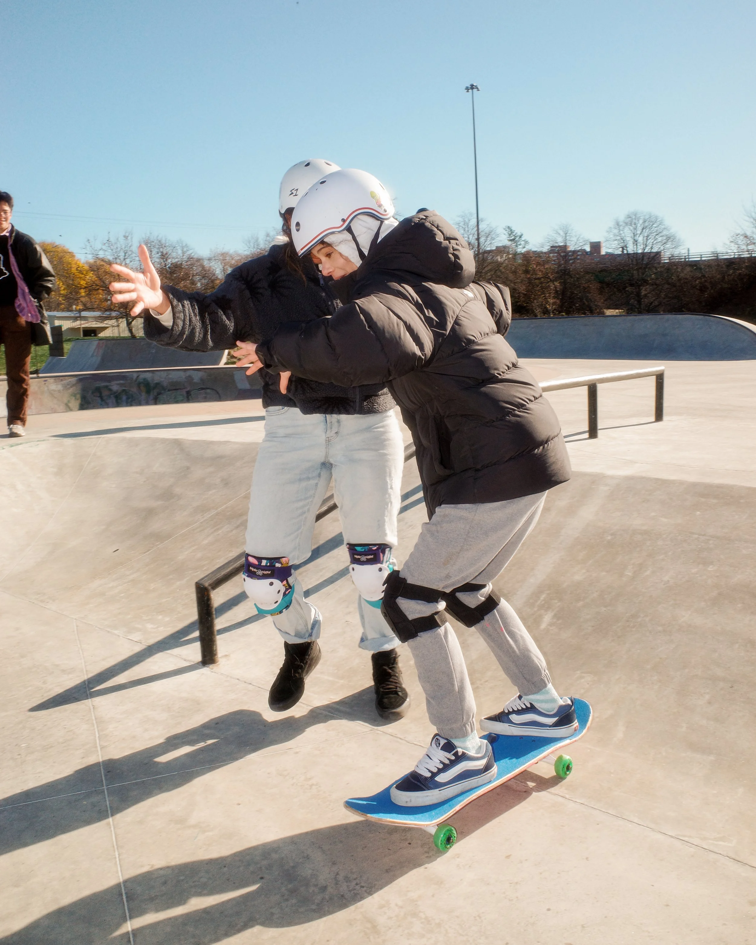 Two people skateboarding at a skate park, one person helping the other with safety gear, both wearing helmets and knee pads, in an outdoor setting during daytime.