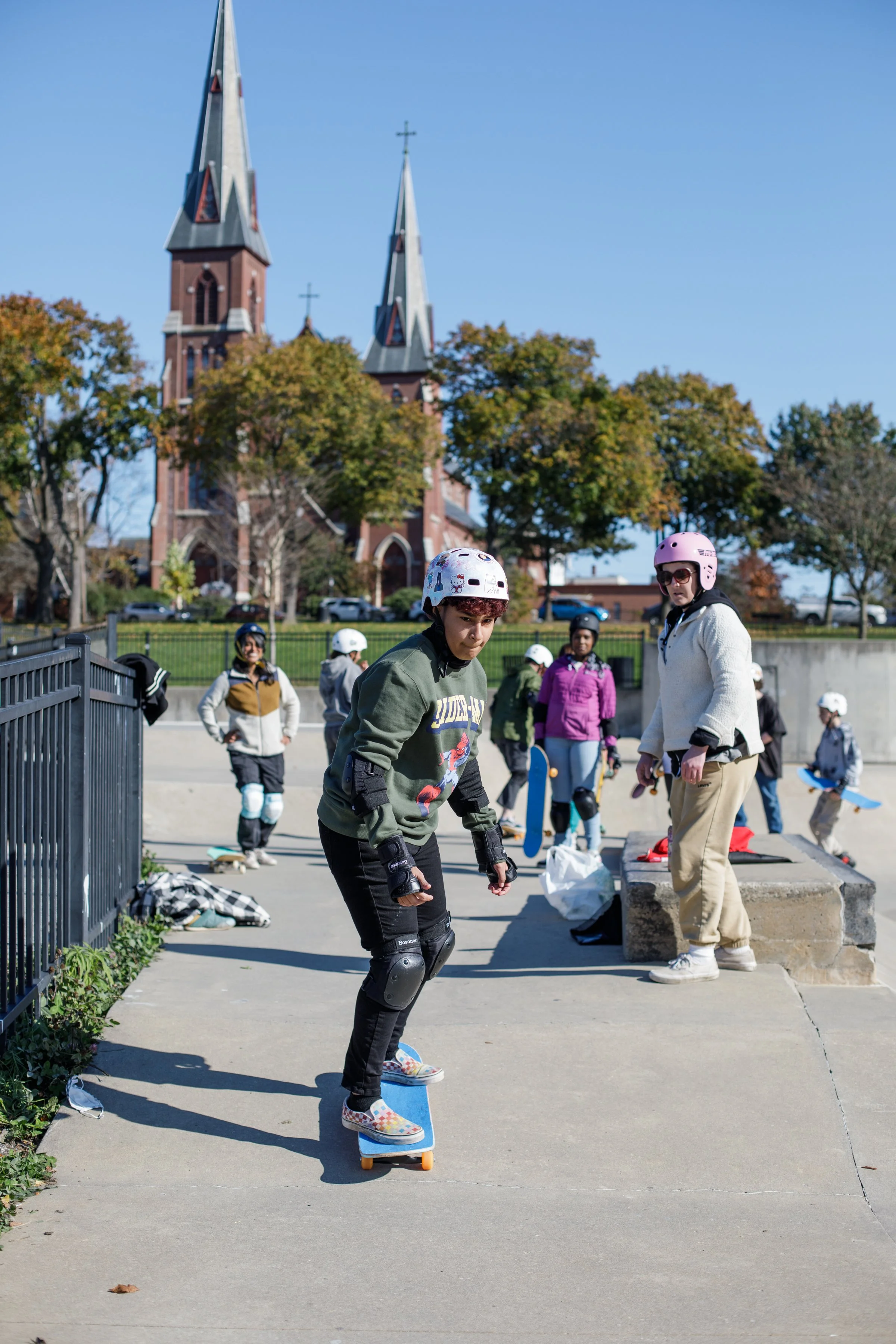 Young person skateboarding at a skate park with others in the background, under a clear blue sky, with a church and trees in the distance.