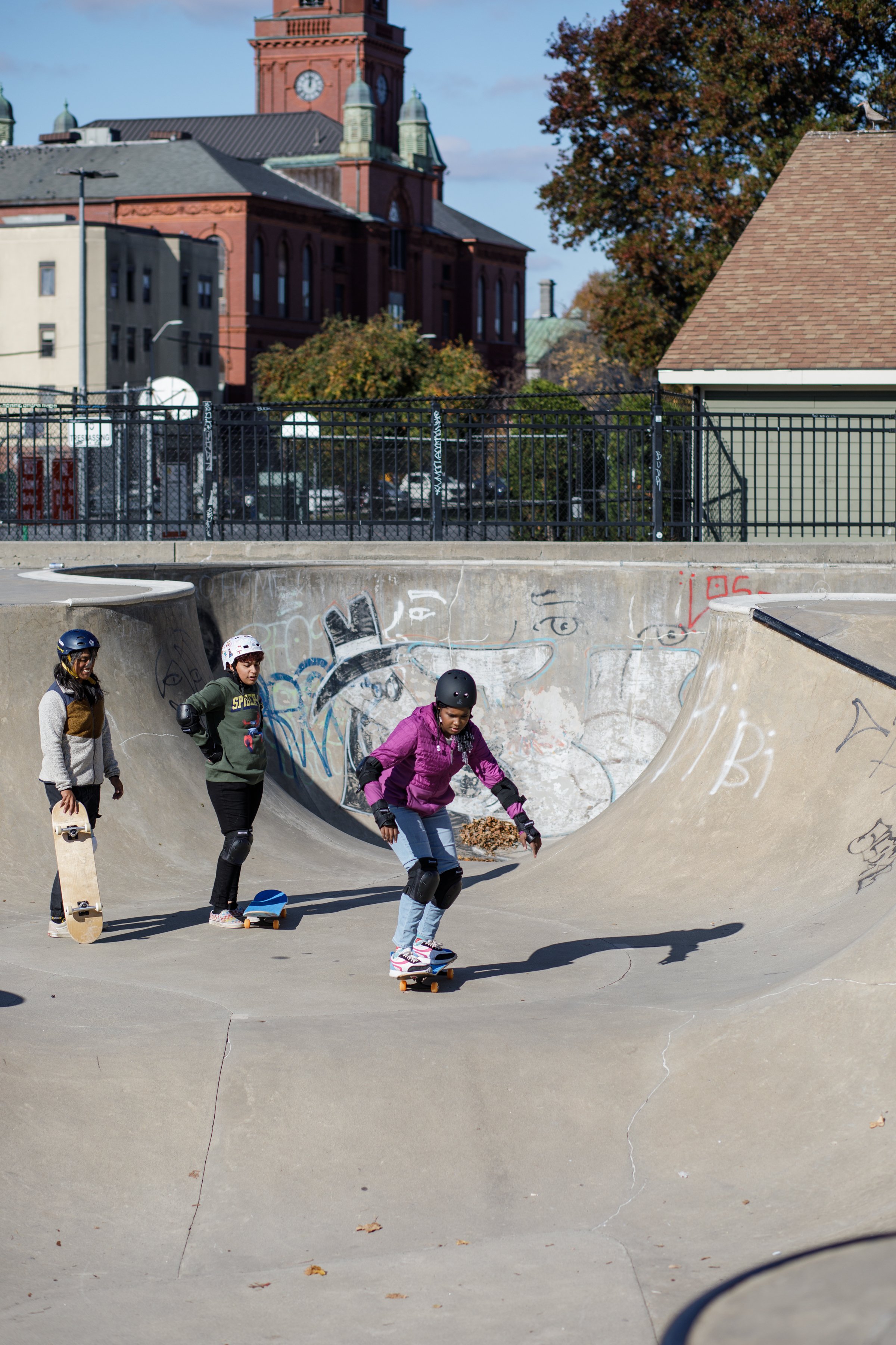 Three young girls skateboarding at a skate park during daytime, with a brick building and trees in the background.