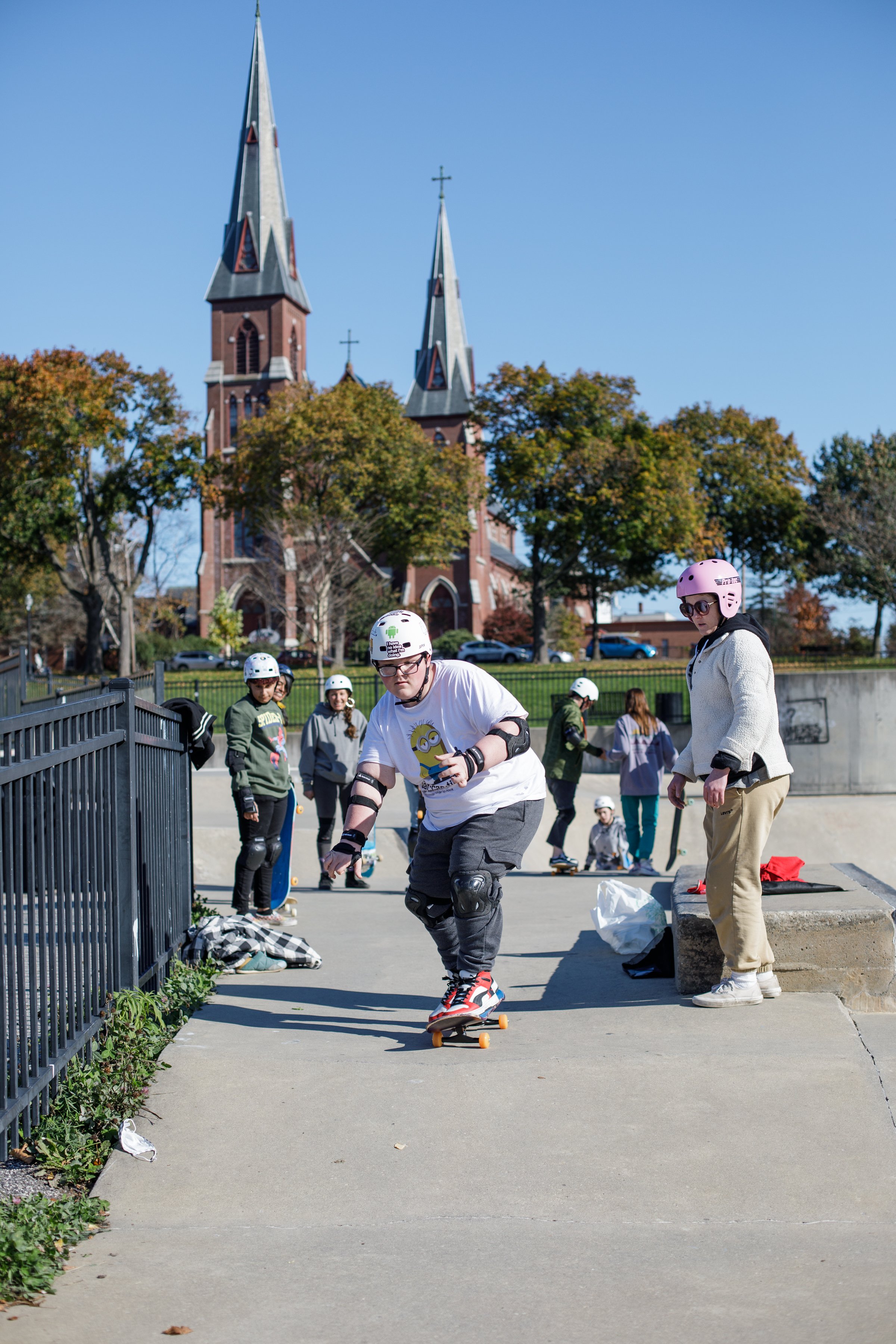 People skateboarding and rollerblading at a skate park on a sunny day, with a church and trees in the background.
