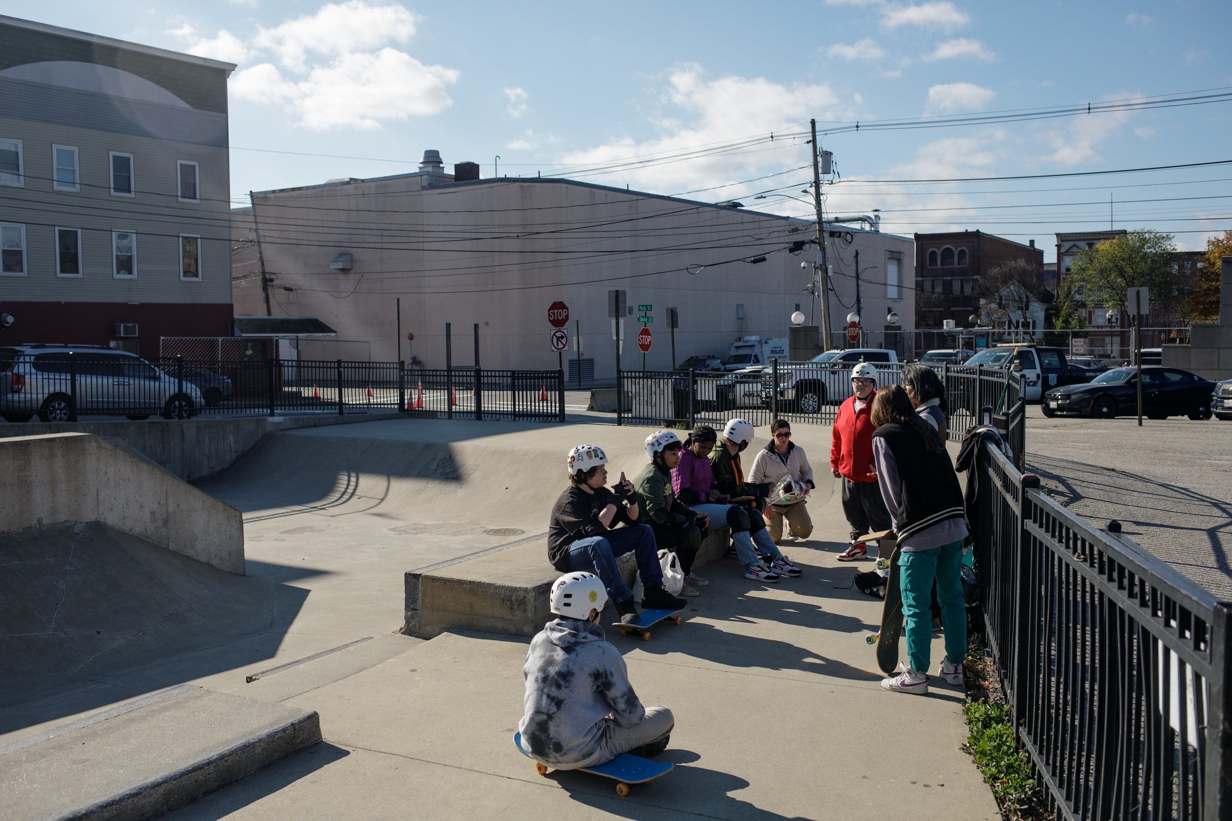 Group of young people with skateboards sitting and standing at a skate park, some wearing helmets, in an urban area on a sunny day.