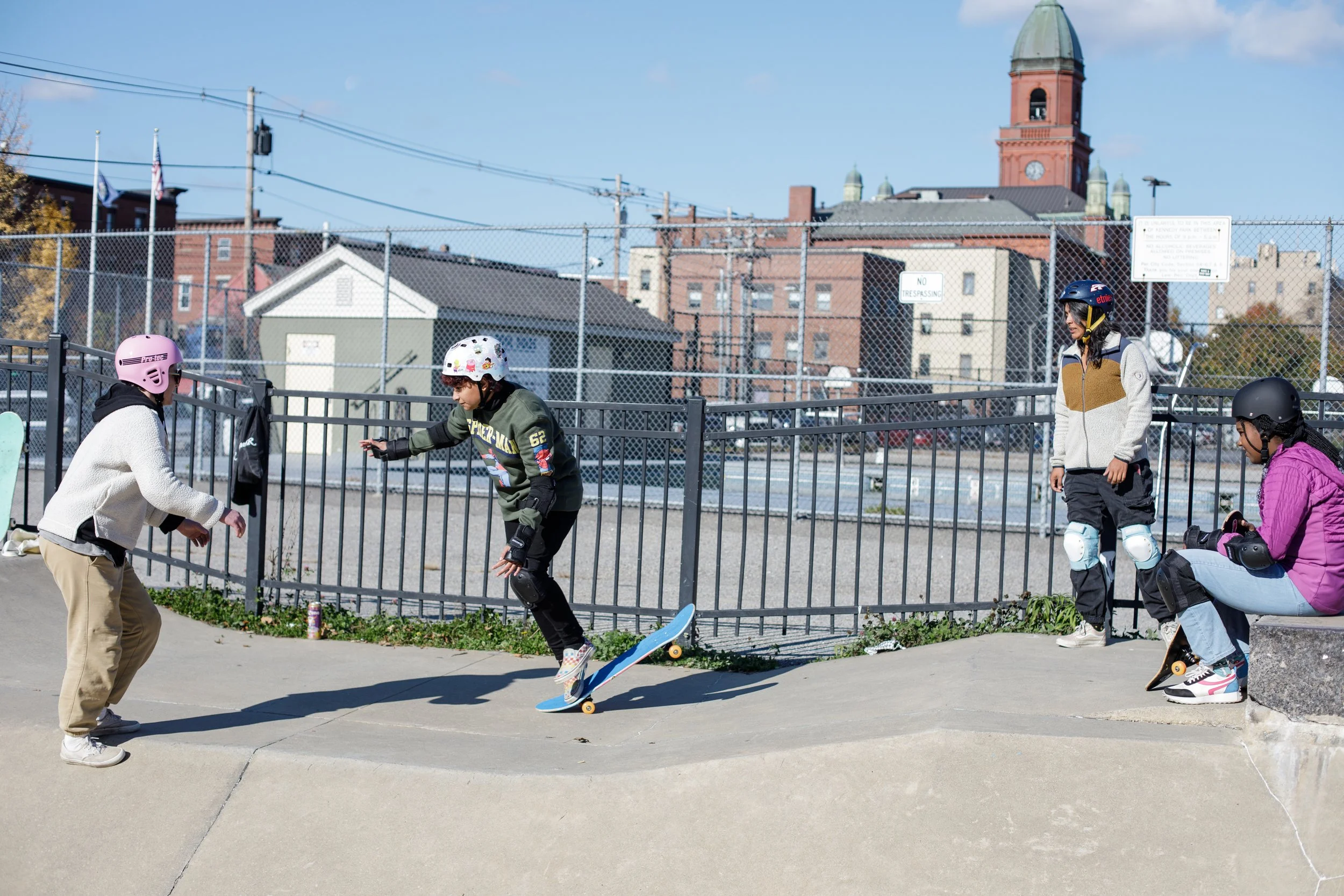 A group of young people at a skate park, with one person skateboarding while others watch or prepare, all wearing helmets and safety gear, with a cityscape and chain-link fence in the background.