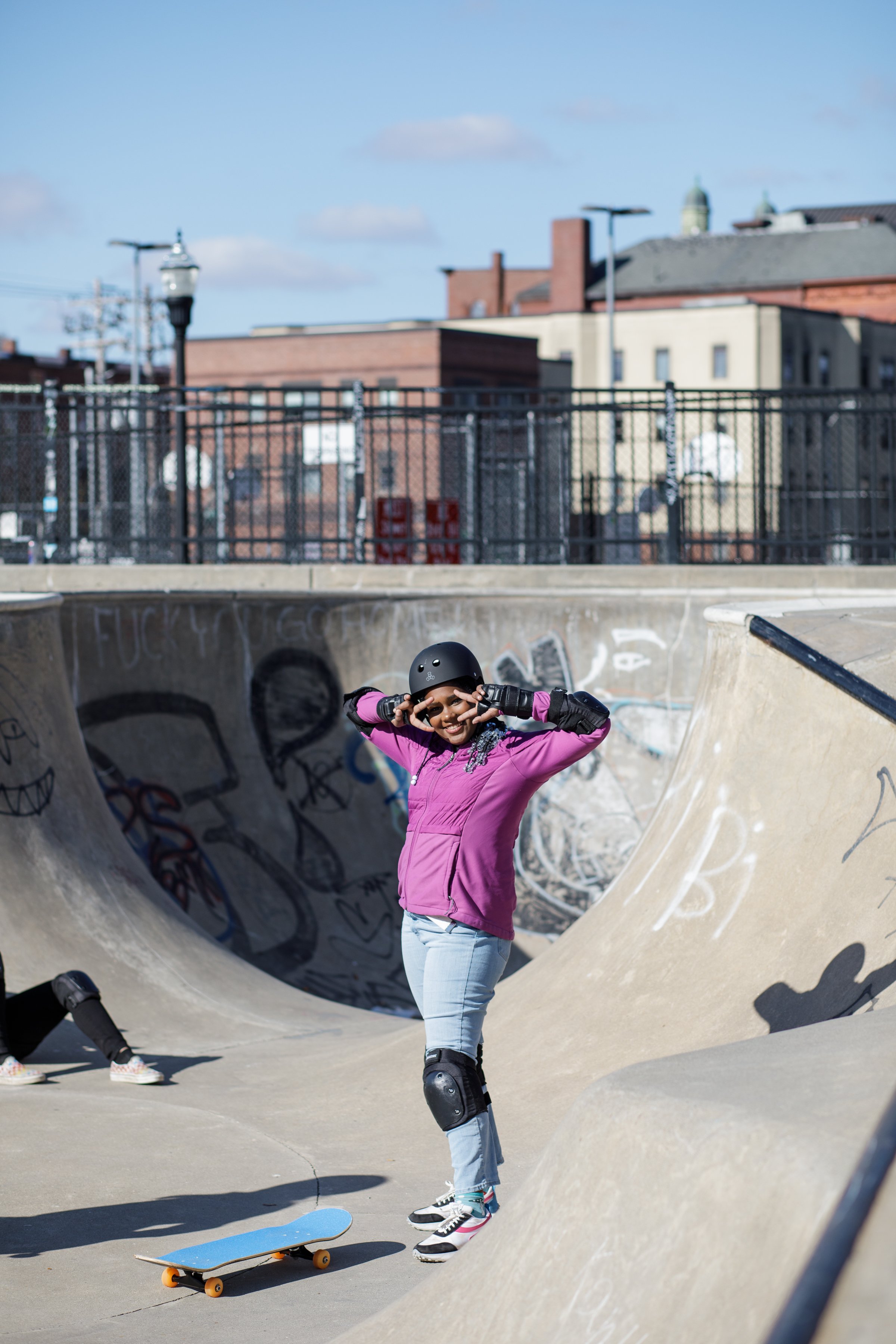 A young girl wearing a pink jacket, helmet, and knee pads, standing at a skate park, smiling and holding her helmet while a skateboard lies on the ground nearby.