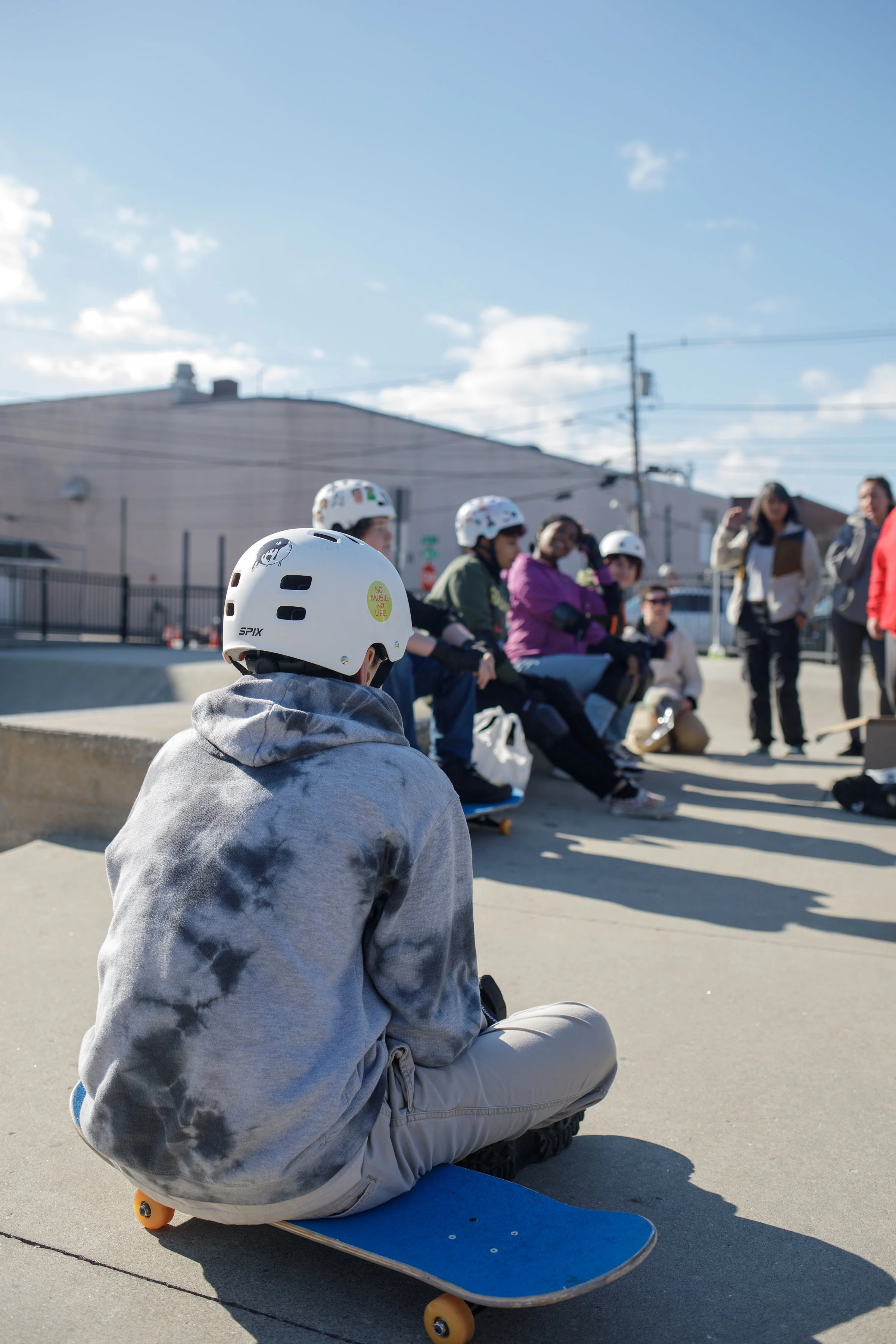 A person sitting on a skateboard wearing a white helmet, at a skate park with a group of people also wearing helmets gathered nearby.