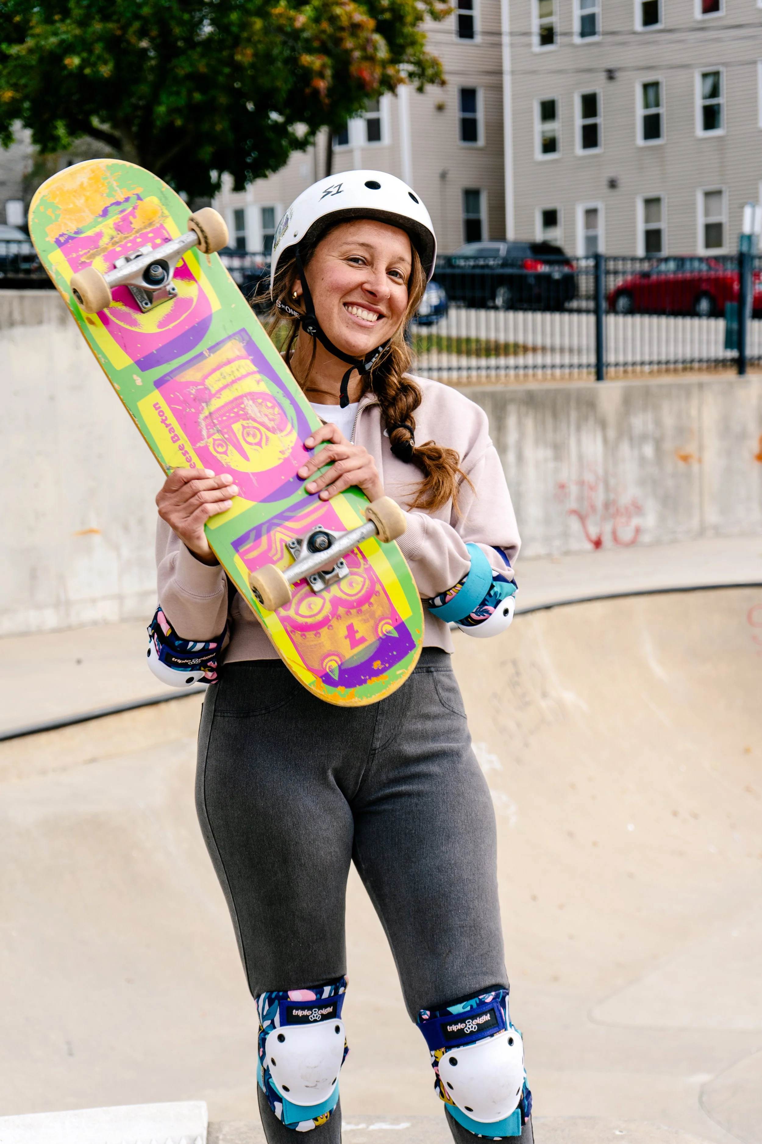 A smiling woman wearing a white helmet, knee and elbow pads, holding a skateboard at a skate park.
