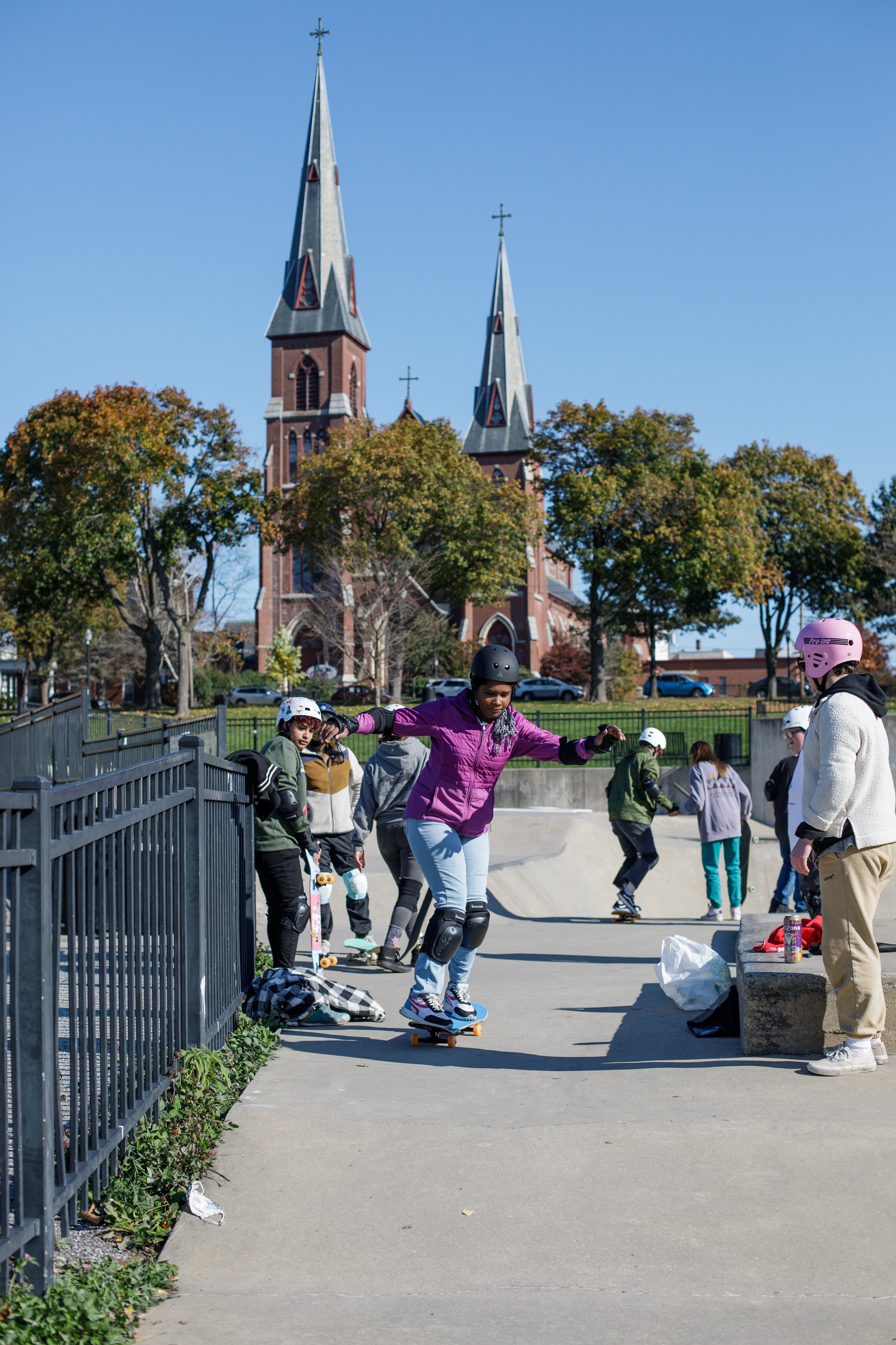 People skating at a skate park with a church and trees in the background on a clear, sunny day.