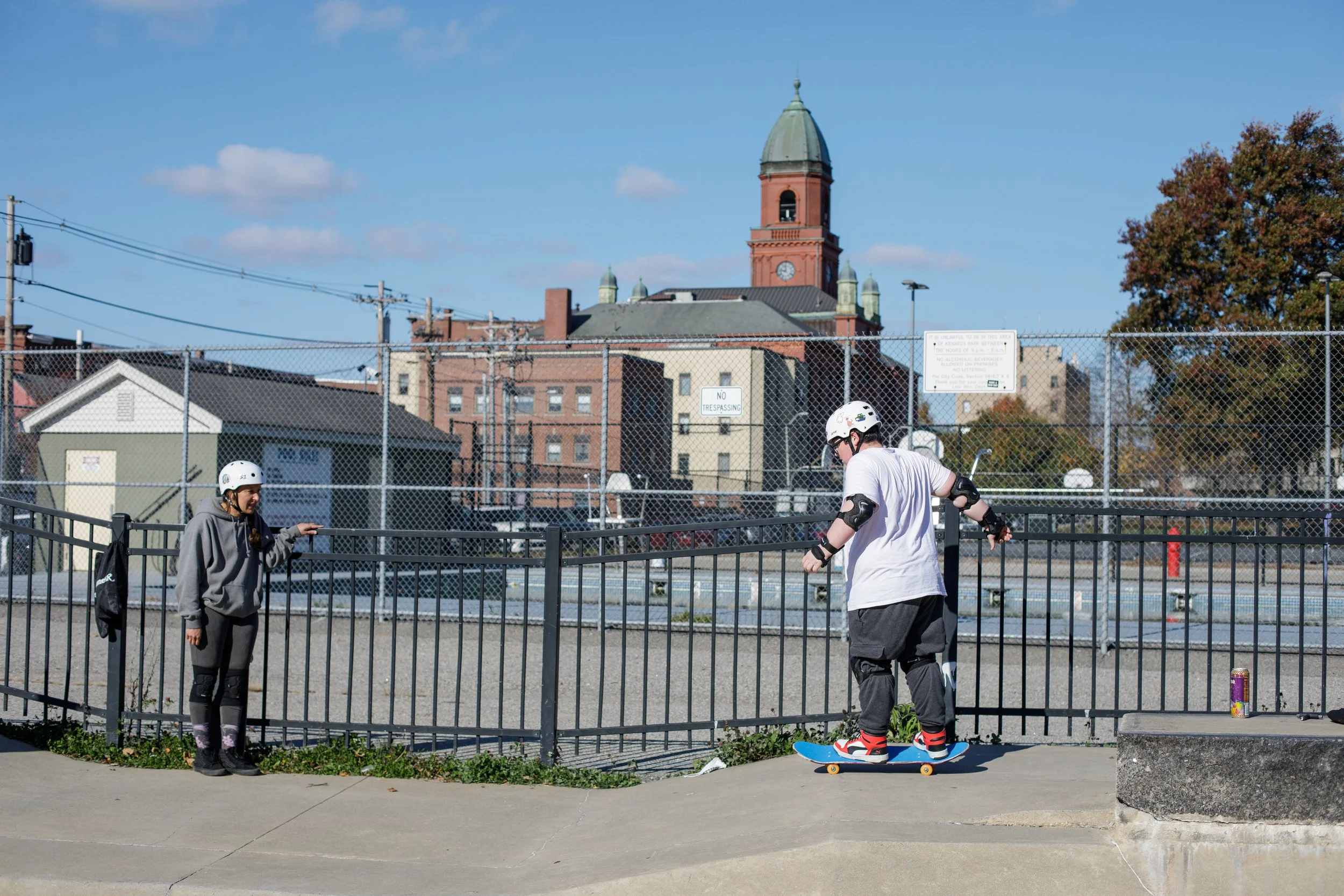 Two skateboarders wearing helmets and protective gear practicing at a skate park, with a cityscape and church tower in the background on a sunny day.