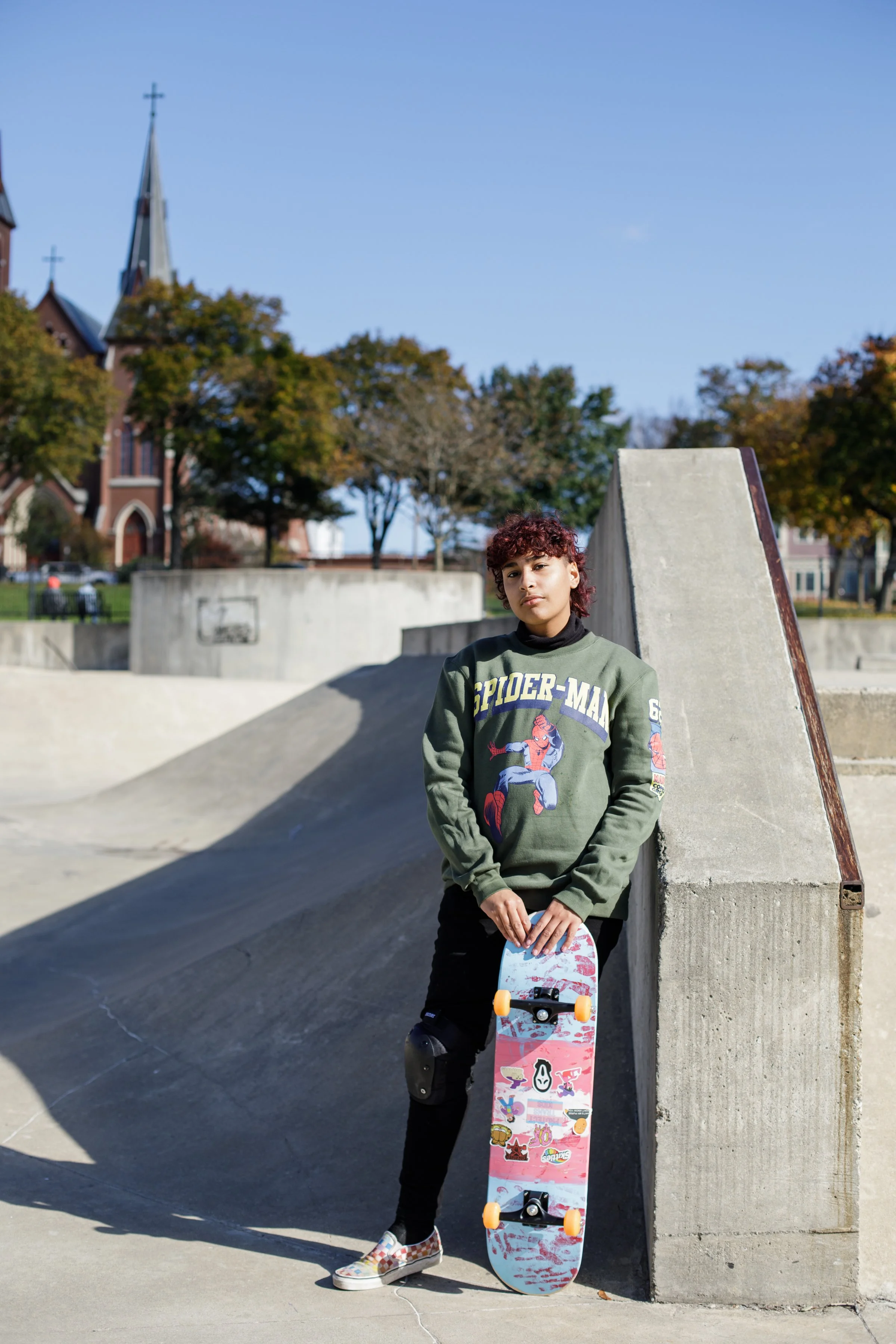 Young person with curly hair in a green Spider-Man sweatshirt and black pants with a skateboard, standing against a concrete ramp at a skate park on a sunny day.