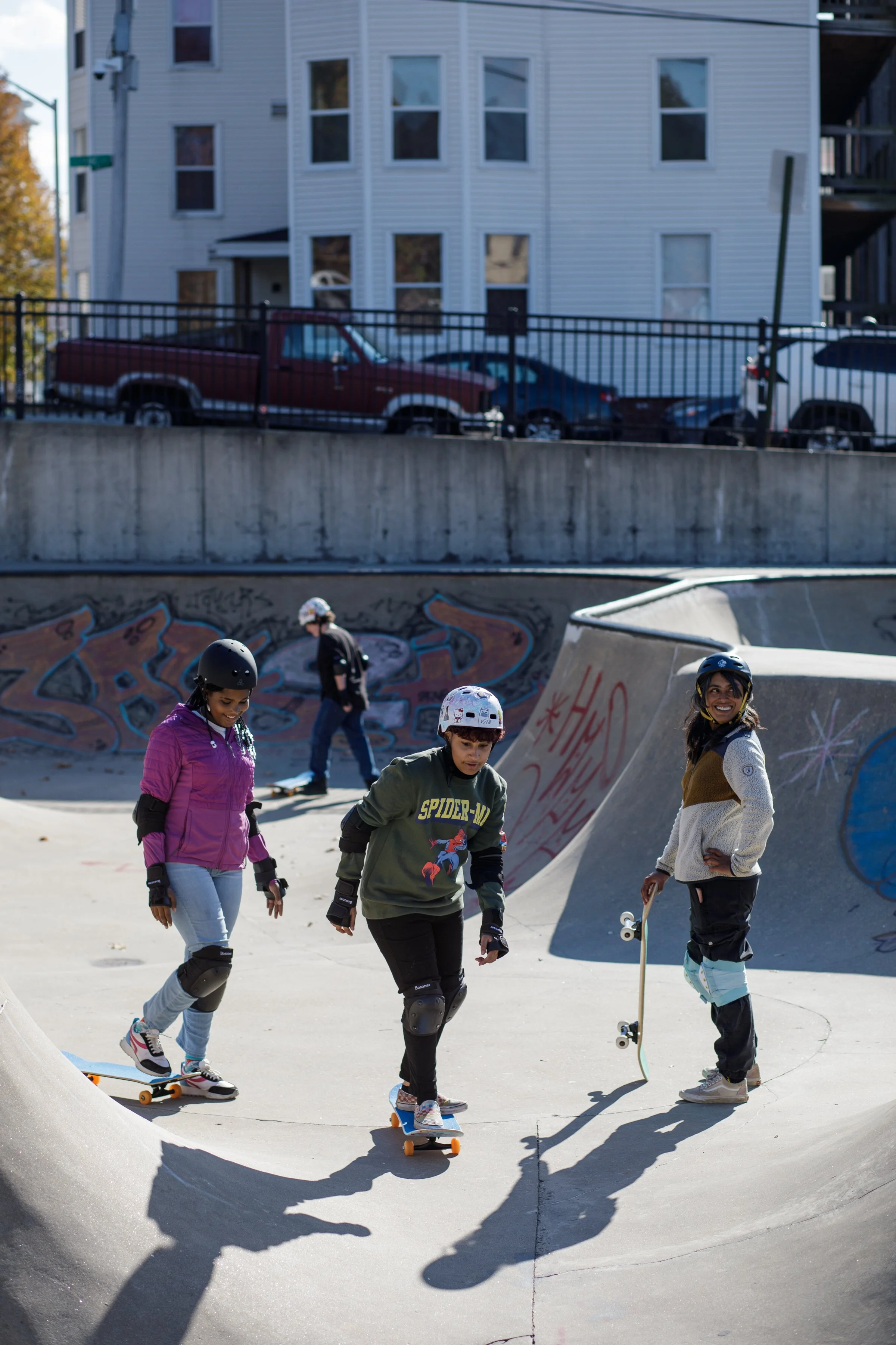 Four kids skateboarding at a skate park, one of them holding a skateboard and smiling, all wearing helmets and safety gear, with graffiti on the concrete ramps.