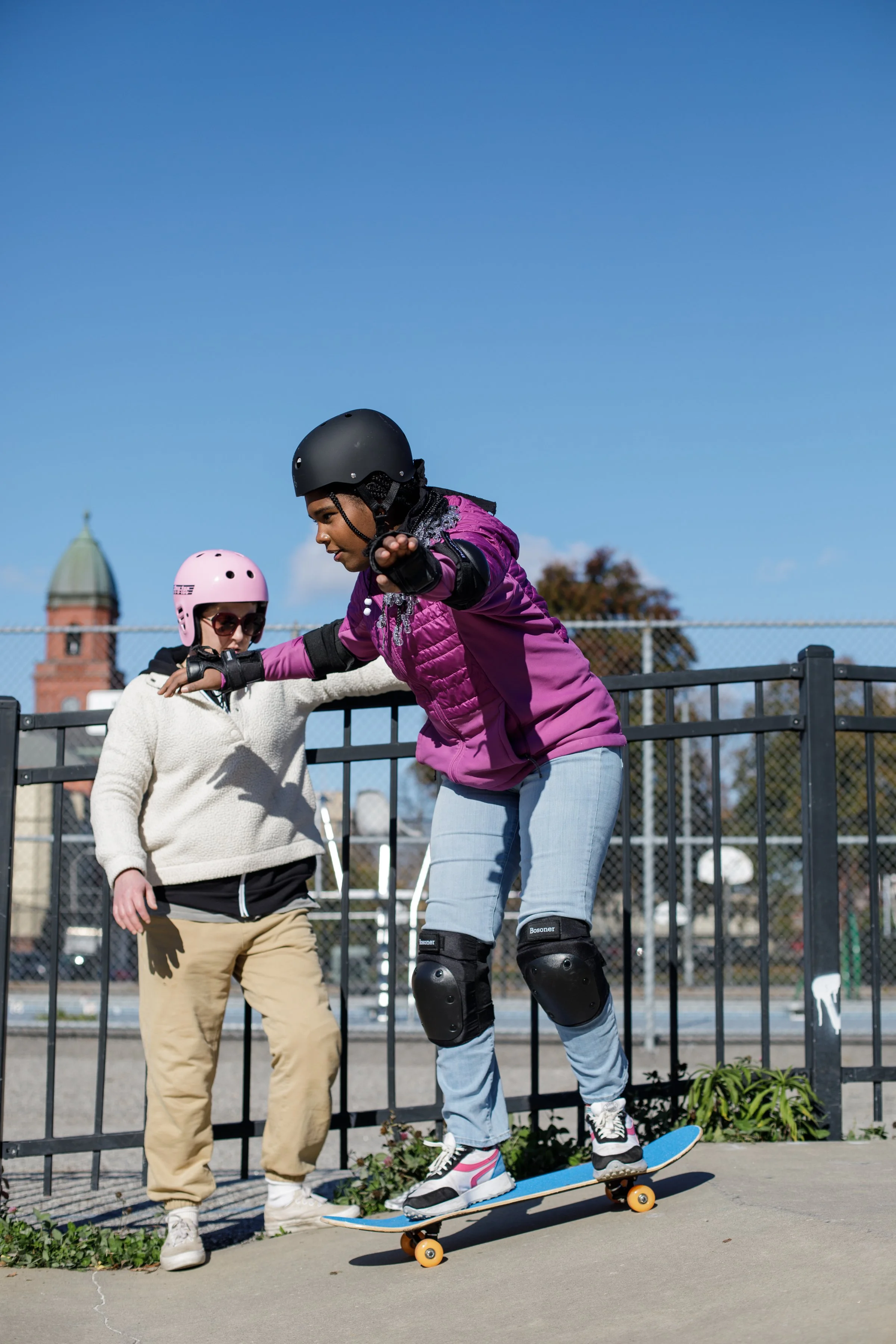 A woman wearing a purple jacket and black helmet skateboarding with a young child in pink helmet and sunglasses at a skate park on a sunny day.