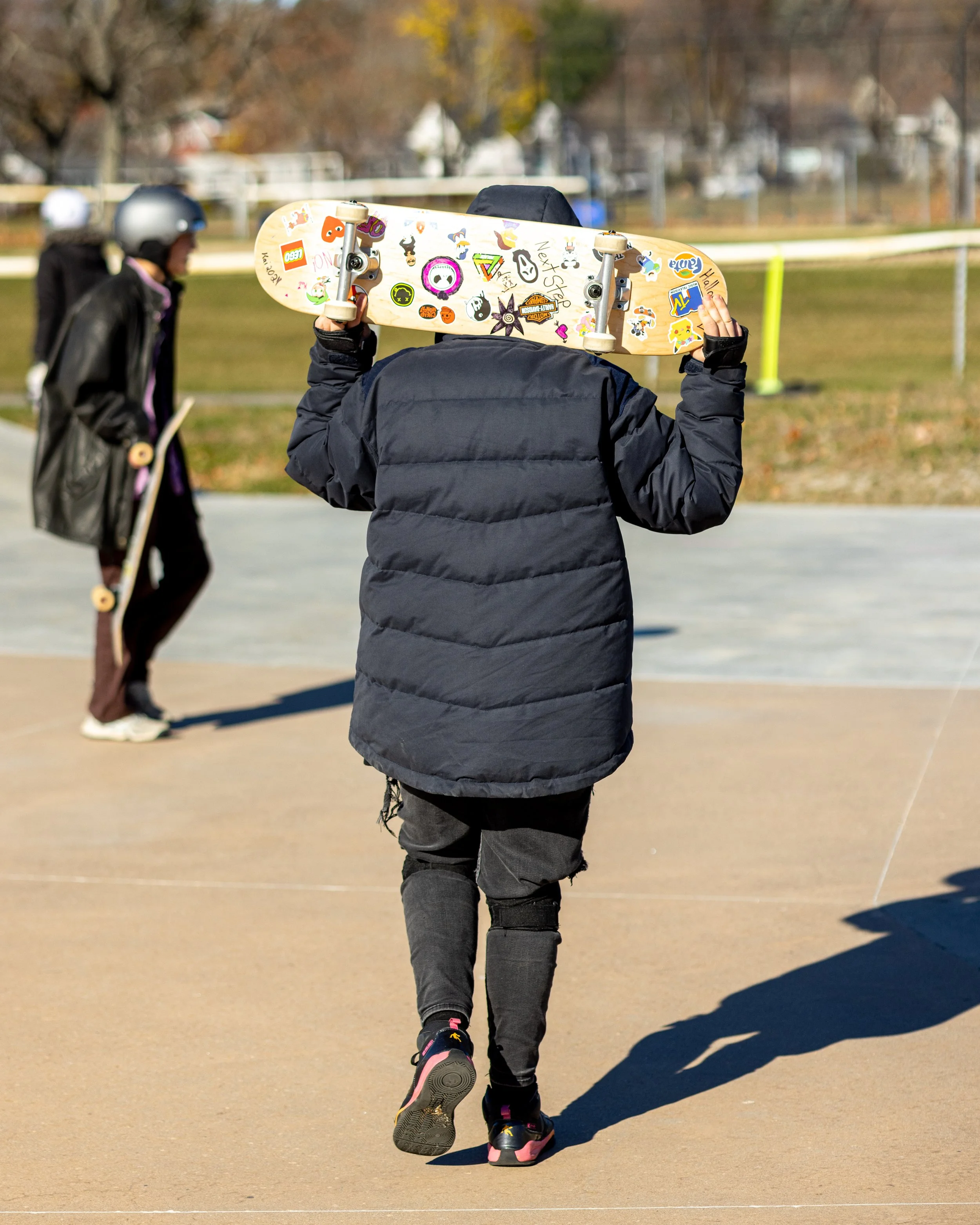 Person in a black puffy jacket carrying a skateboard with colorful stickers over their shoulder at a skate park on a sunny day, with another person and trees in the background.
