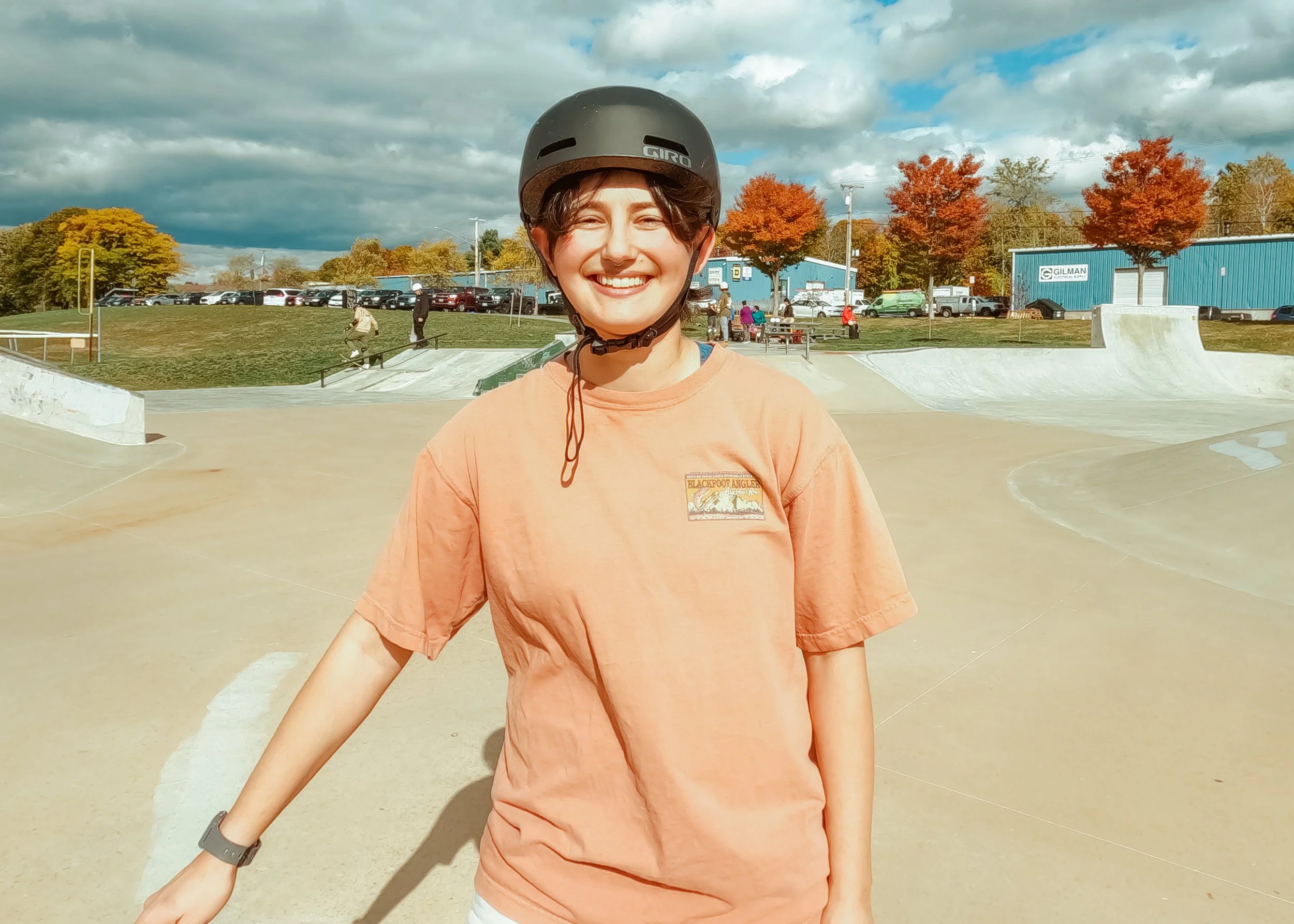 Young woman in an orange t-shirt and helmet smiling at a skate park with autumn trees and parked cars in the background.