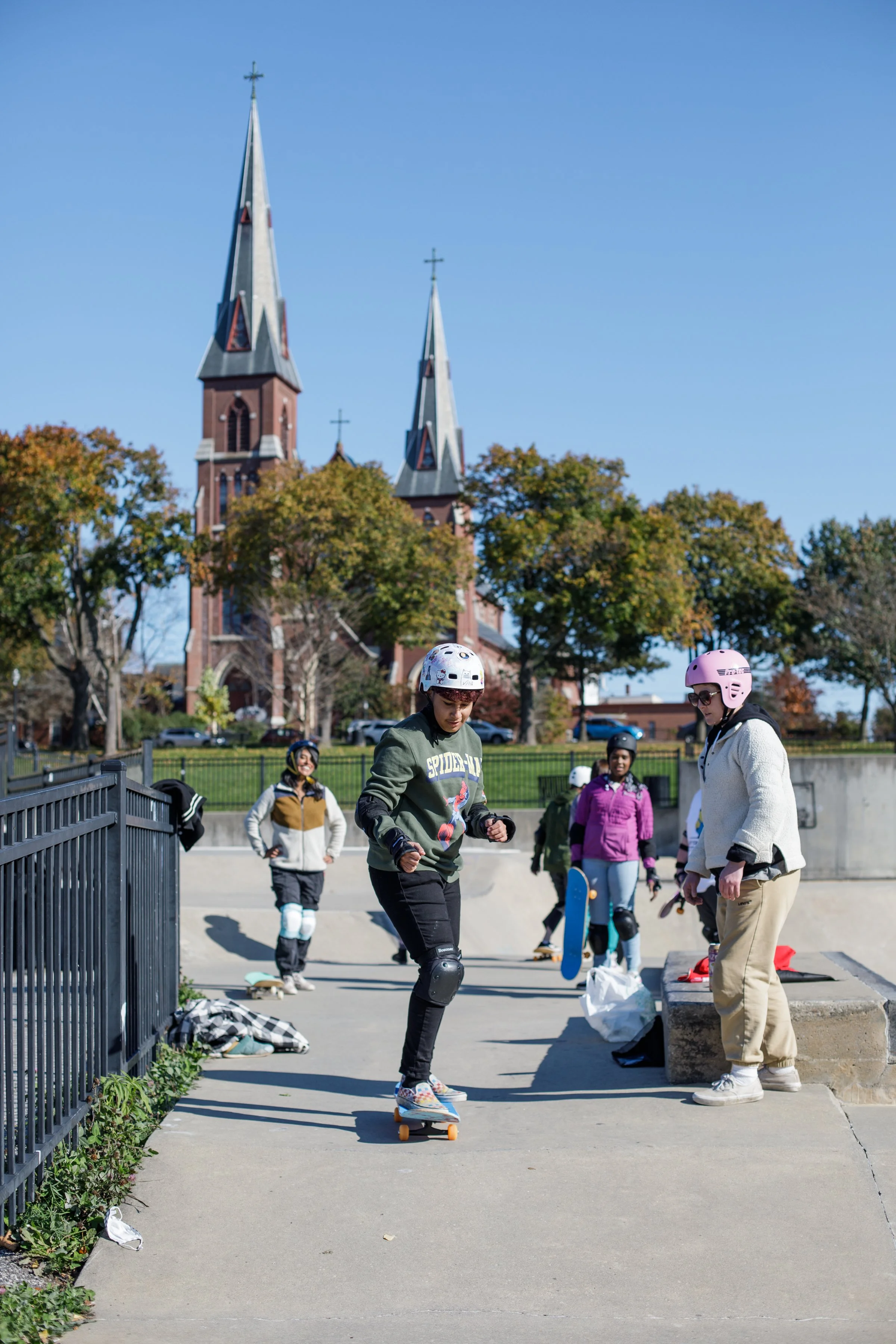 Group of young skaters at a skate park with a church and trees in the background on a clear, sunny day.