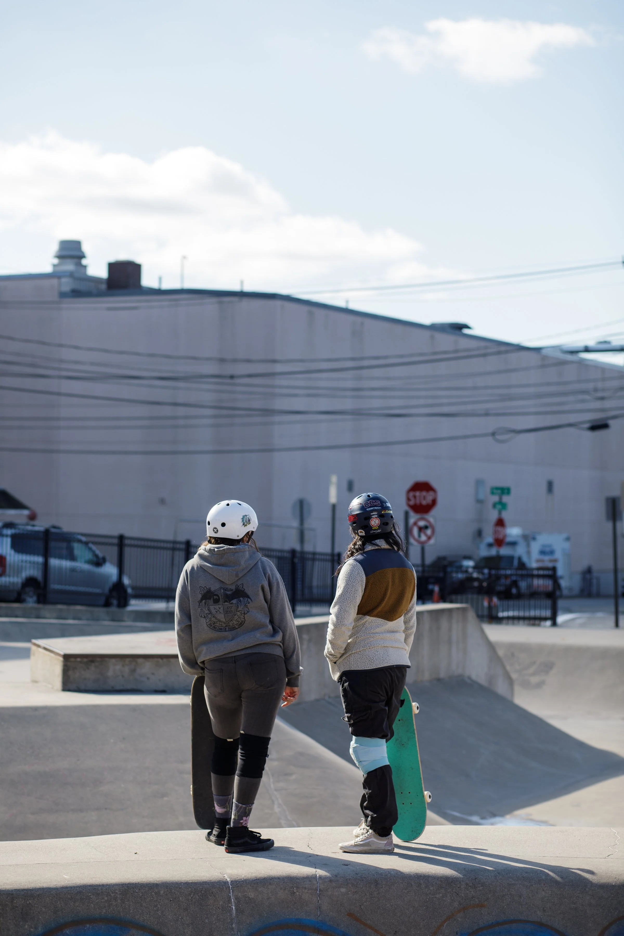 Two skateboarders, wearing helmets, standing on a concrete ramp at a skate park, with one holding a skateboard, under a partly cloudy sky.