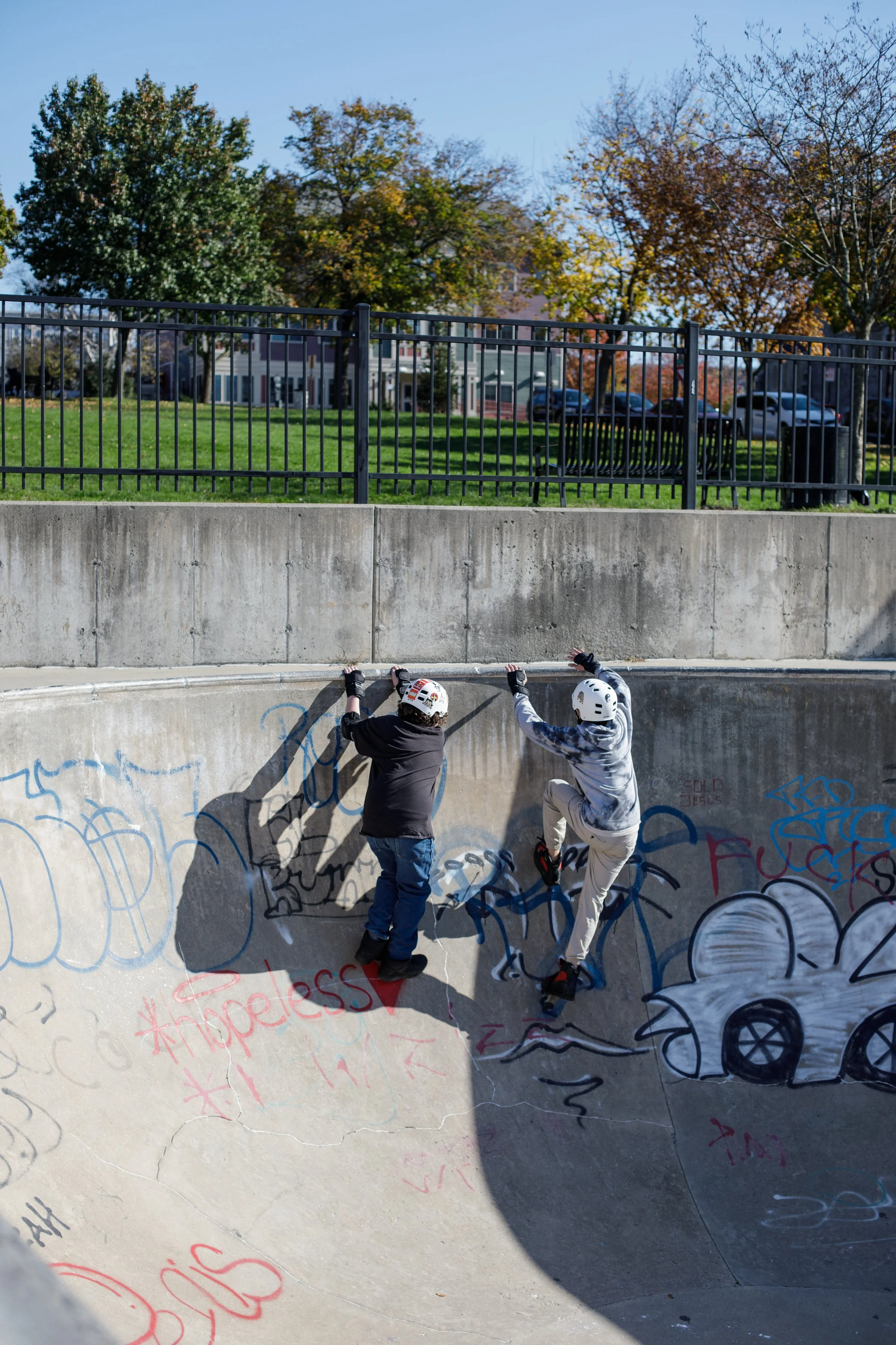 Two people wearing helmets and gloves are climbing the wall of a concrete skate park bowl decorated with graffiti, with a park and trees in the background.