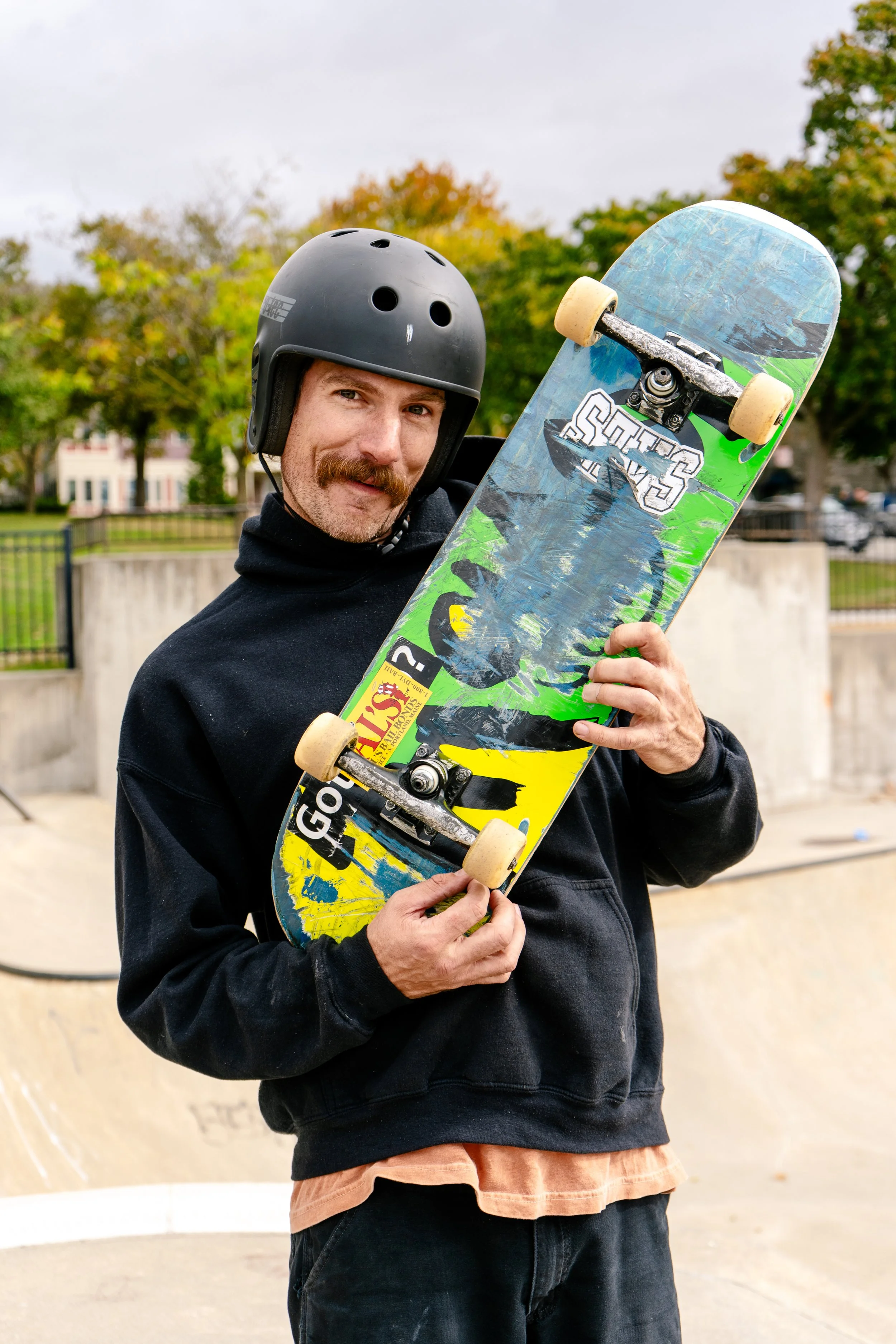 A man wearing a black helmet and a black hoodie holding a skateboard in a skate park with trees and a fence in the background.