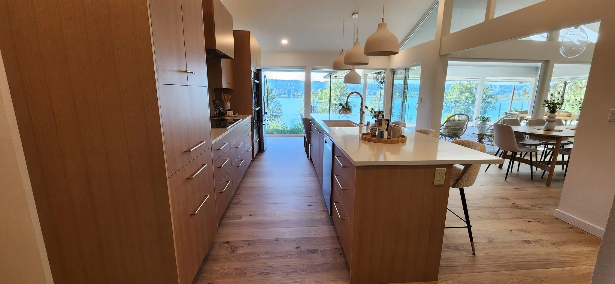 Modern kitchen with a white countertop island, wooden cabinets, and large windows overlooking a lake and trees outside.