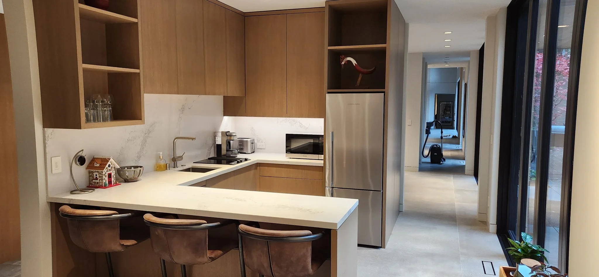 Modern kitchen with wooden cabinets, white countertops, stainless steel appliances, and a breakfast bar with three chairs. There is a microwave, toaster, coffee maker, and a small house-shaped decorative item on the counter, with open shelves displaying glasses and a sculpture. A hallway leads to another room, with a vacuum cleaner visible in the distance. Large windows let in natural light.