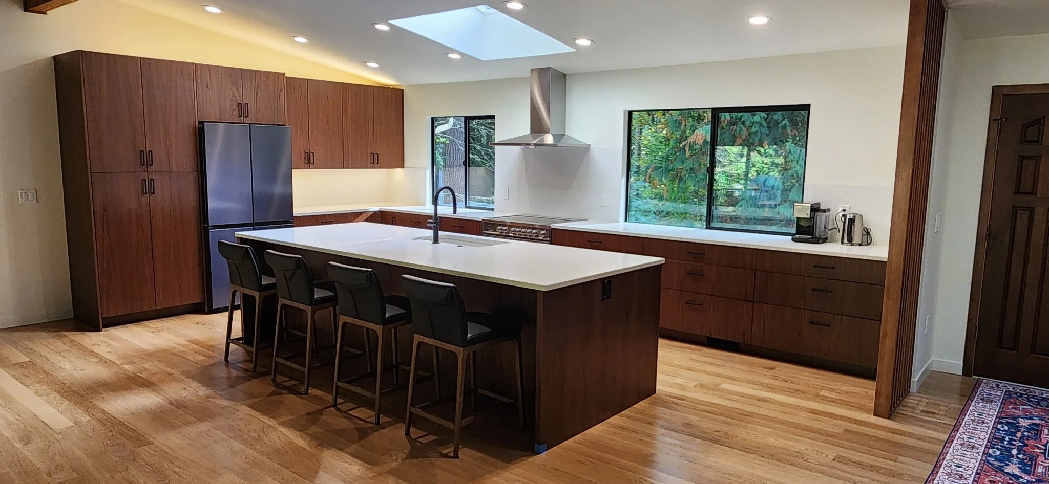 Modern kitchen with wooden cabinets, white countertops, and hardwood floors. Features an island with four black chairs, a stainless-steel refrigerator, a stove with a vent hood, and large windows overlooking a lush green outdoor view.
