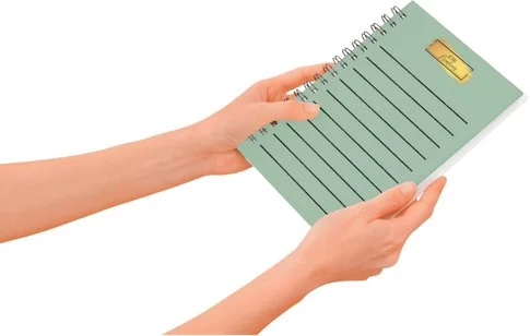 A person's hands holding a green spiral-bound notebook with a gold thumbtack on the cover.