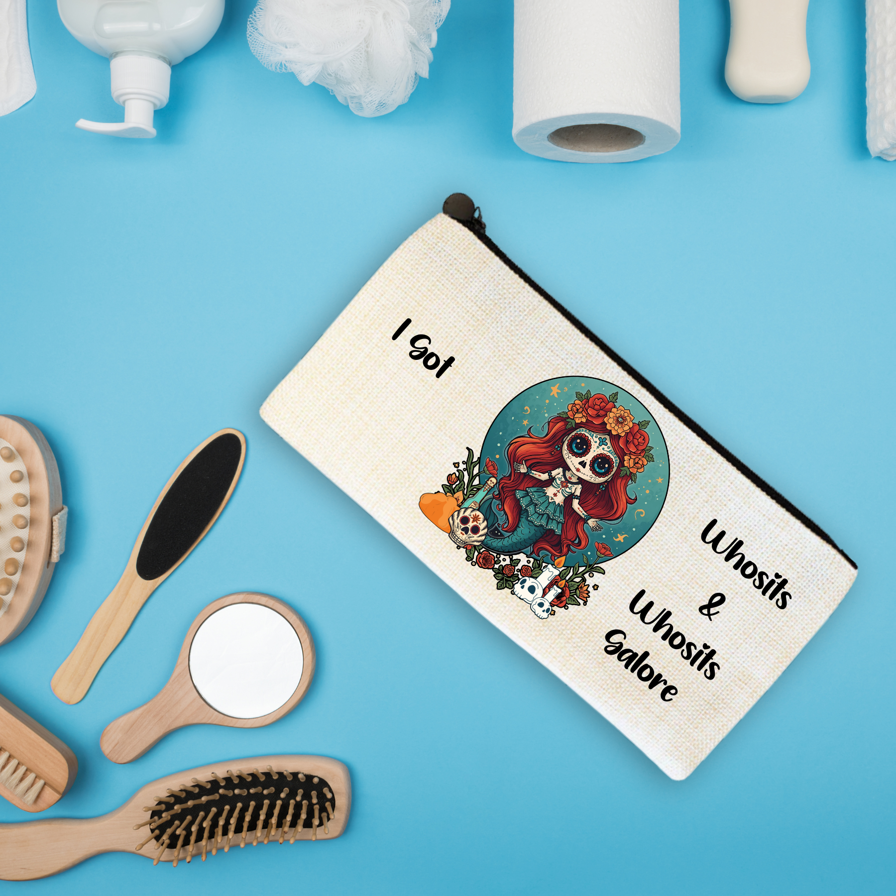A toiletry bag with the text "I got Unsits & Unosit Carore" and an illustration of a girl with red hair, flowers, sugar skull face paint, surrounded by smaller skulls and flowers, on a blue background. Various grooming items such as brushes, a mirror