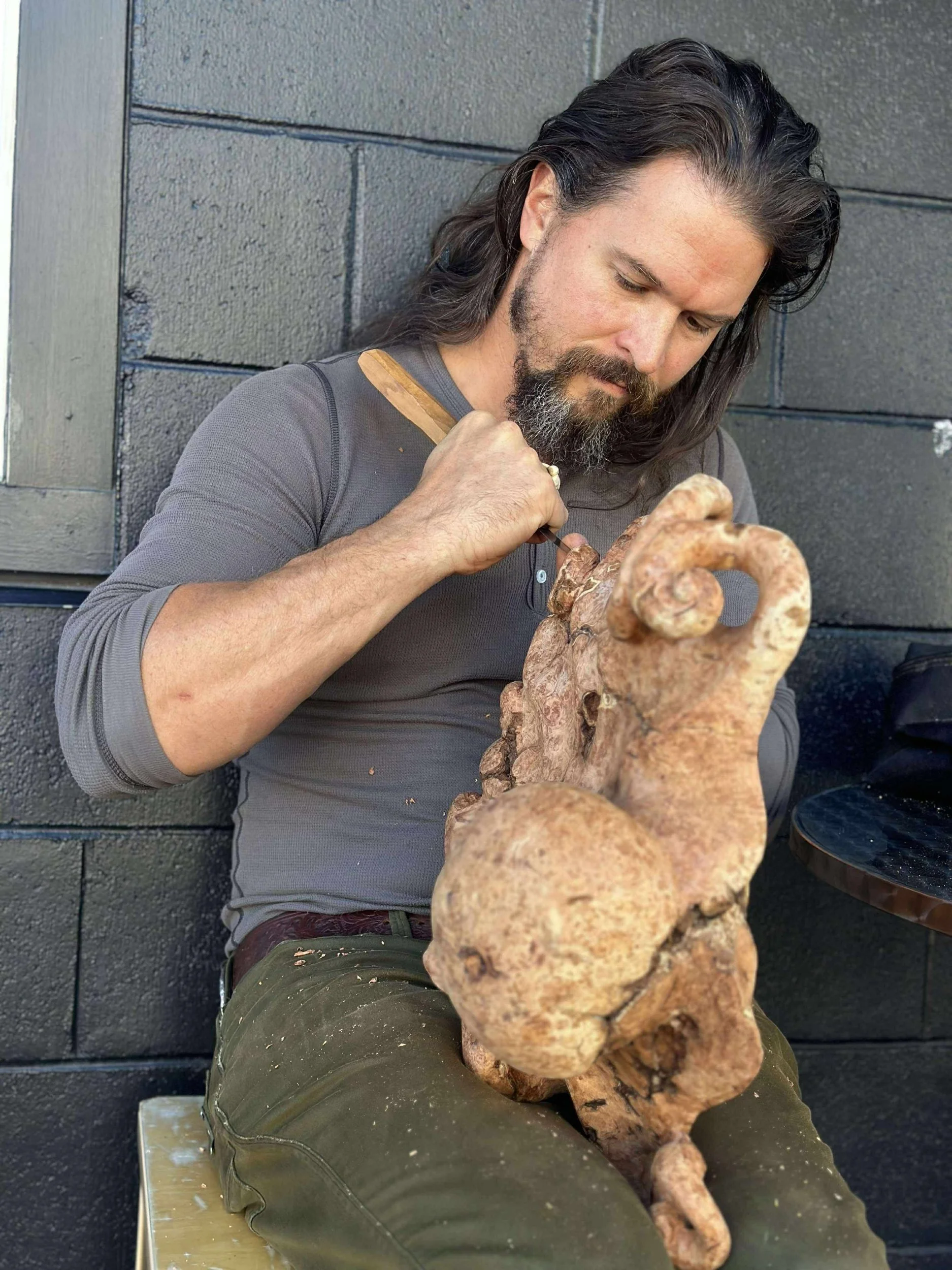 A man with long dark hair and a beard is sitting outdoors against a dark gray wall, intently carving a large, gnarled piece of wood or root.