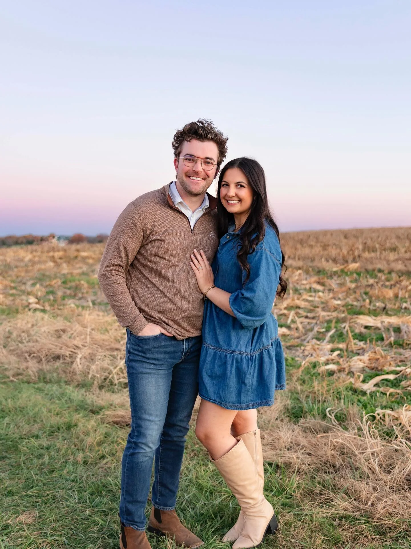 A happy couple stands outdoors in a field with a sunset sky in the background. The man has curly brown hair, glasses, and is wearing a brown sweater and jeans. The woman has long dark hair, and is wearing a blue dress and beige knee-high boots. They are smiling and embracing each other.