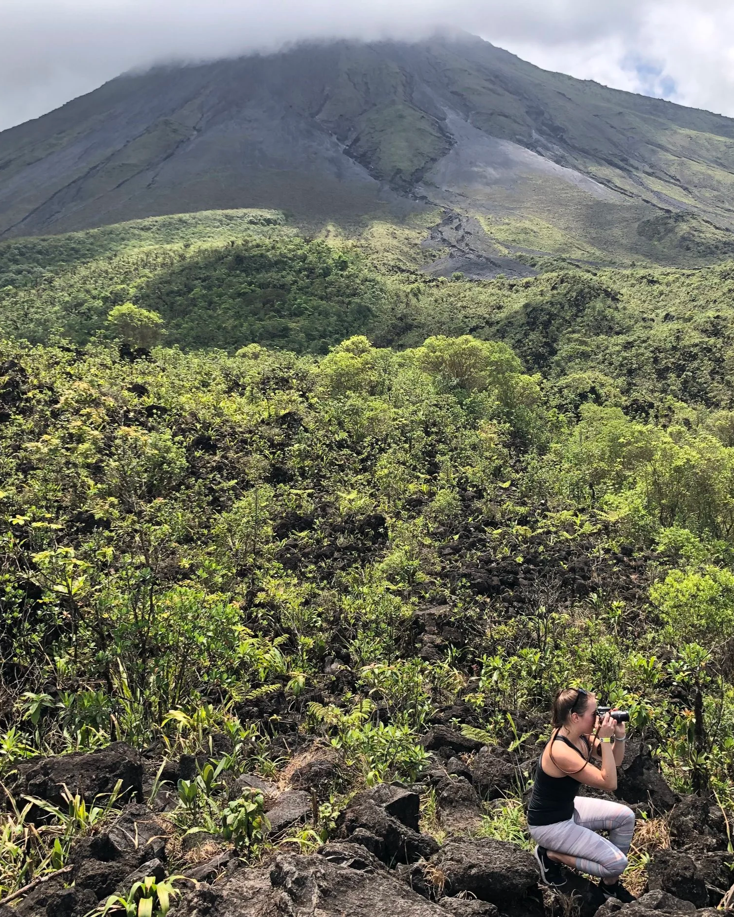 A woman kneeling on rocky terrain in Costa Rica taking photographs of lush green vegetation and a volcano in the background.
