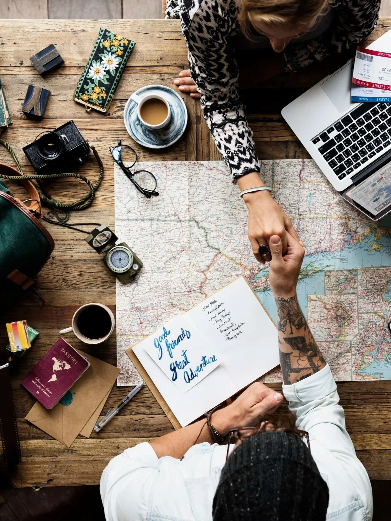 Top-down view of a wooden table covered with travel items, including a map, a notebook with handwritten notes, a passport, camera, glasses, coffee, a cup of tea, a wristwatch, luggage tags, an open laptop, and a woman's hand shaking hands with another person.