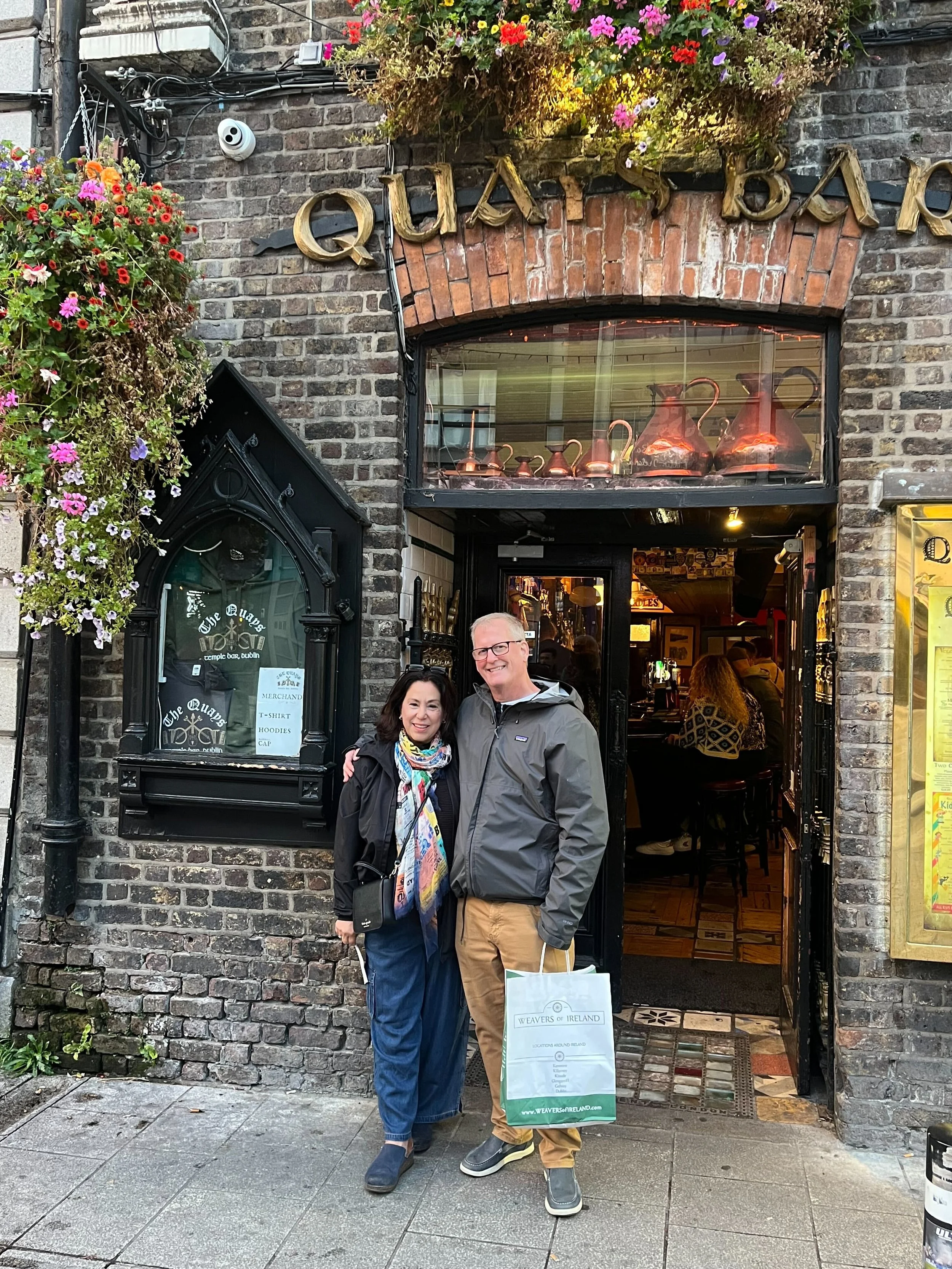 Two people standing in front of The Quays Dublin bar, smiling, with a hanging flower basket above the entrance. The woman is wearing a black jacket, colorful scarf, and jeans, and the man is in a gray jacket and khaki pants holding a shopping bag.