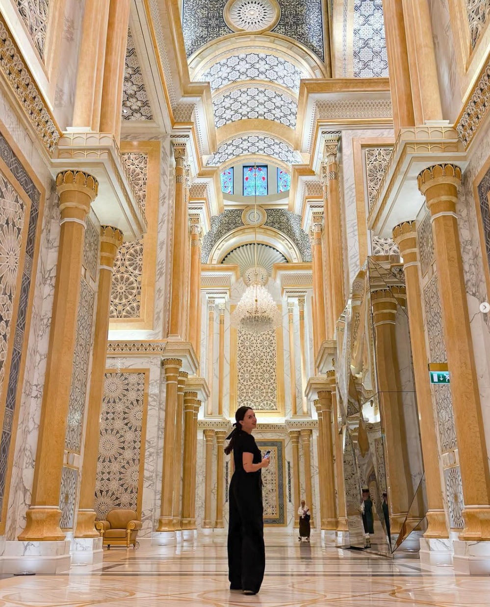 Interior of a luxurious building in the Middle East with tall marble columns, intricate decorative patterns, chandeliers, and stained glass windows, featuring a woman in black looking at her phone.