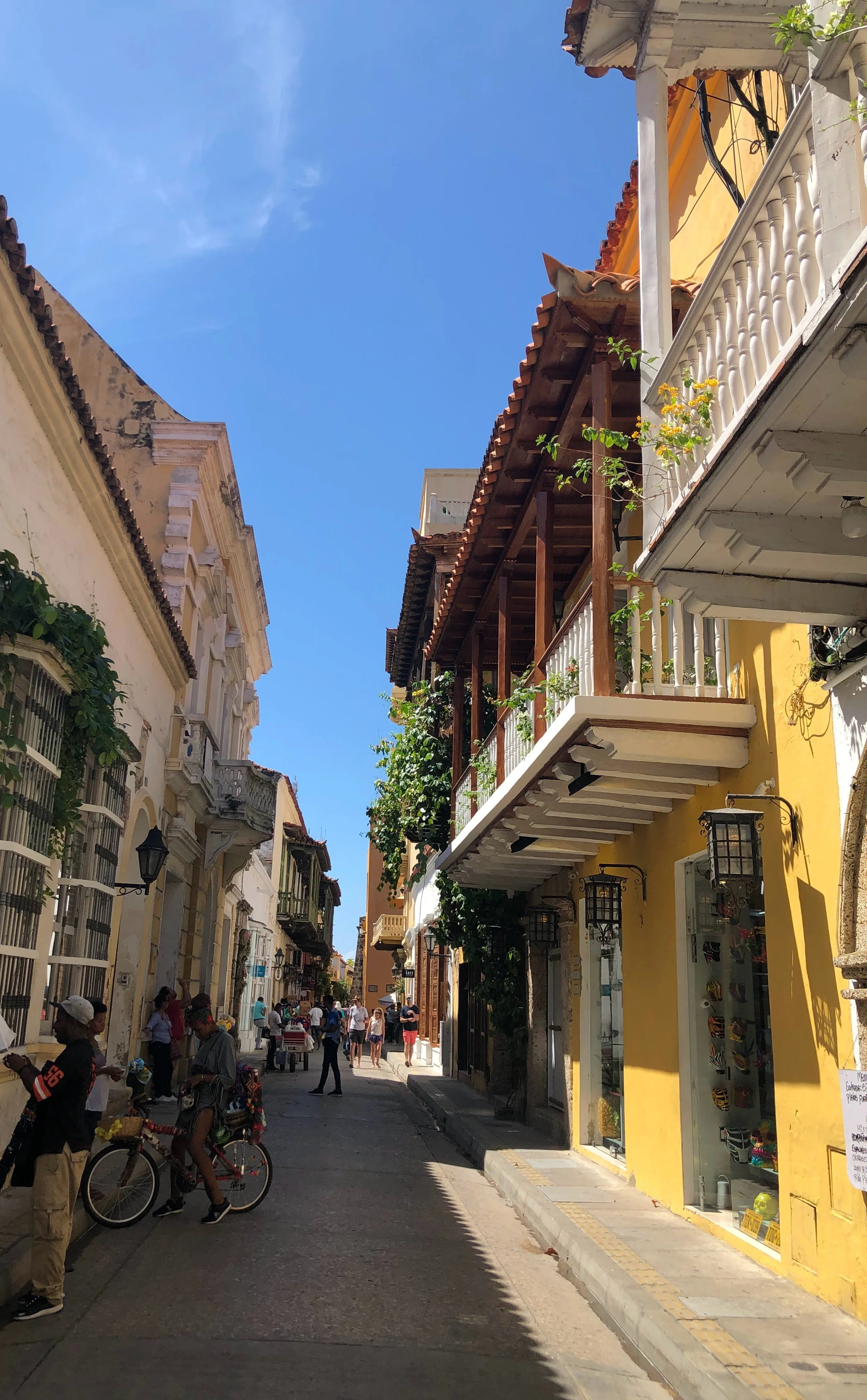 A lively street scene in Colombia with colorful colonial-style buildings, some with balconies, on a sunny day with a clear blue sky. People are walking, and one person is riding a bicycle.