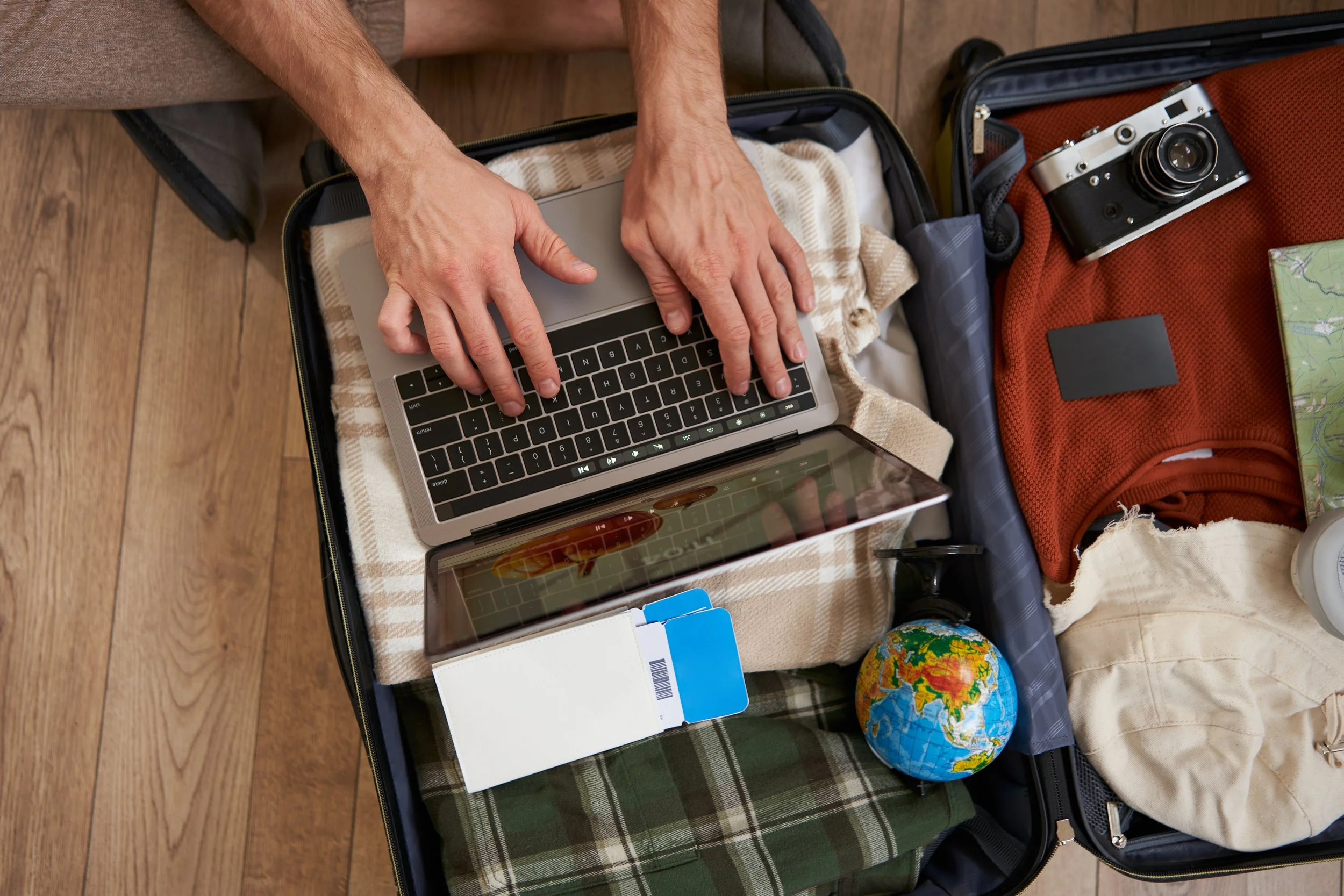 An open suitcase with packing items including a laptop, a tablet, a camera, a globe, a notebook, and clothes on a wooden floor. A person is packing the suitcase.