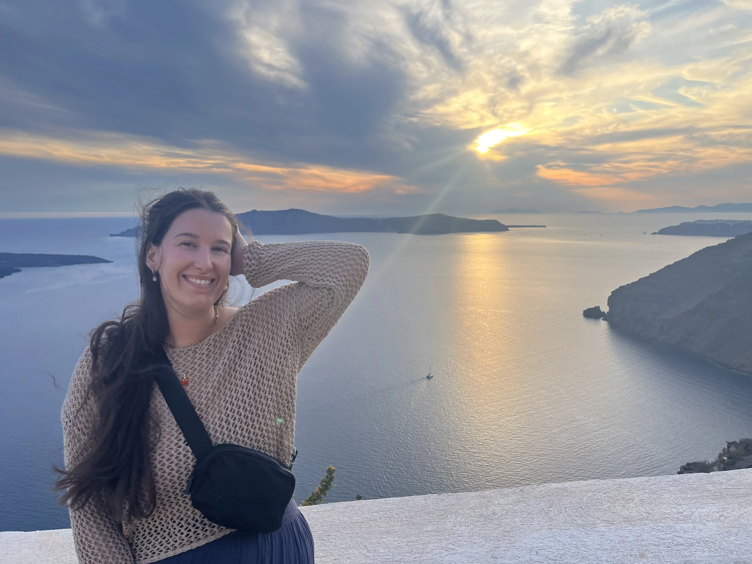A woman with long dark hair smiling and wearing a beige knit sweater, carrying a small black crossbody bag, standing on a white ledge in Greece overlooking the ocean at sunset with mountains and a boat in the distance.