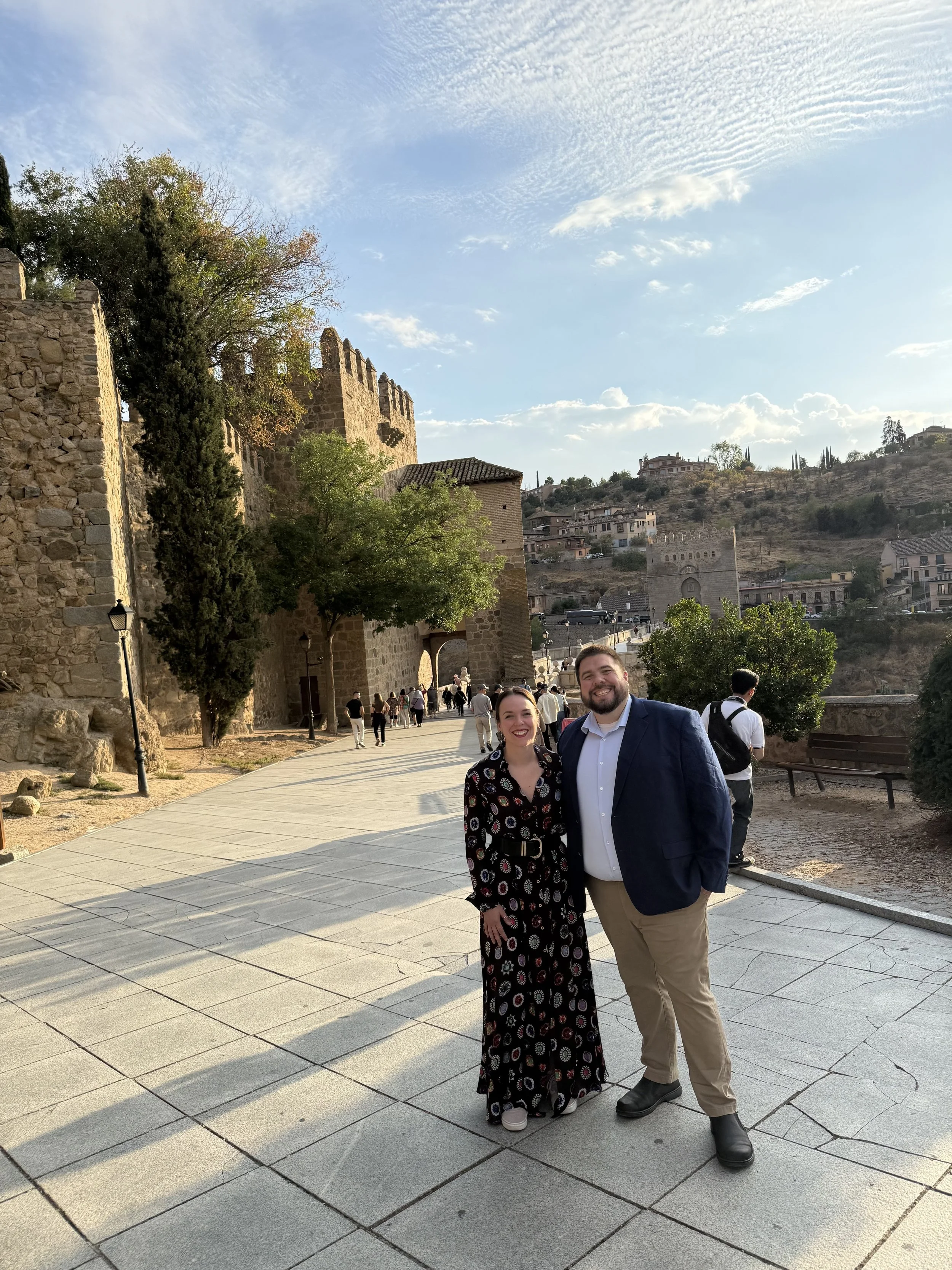 A smiling woman in a black patterned dress and a smiling man in a white shirt and dark blazer pose together outdoors in Spain in front of a historic stone wall with a castle-like structure, trees, and a hillside on a sunny day.