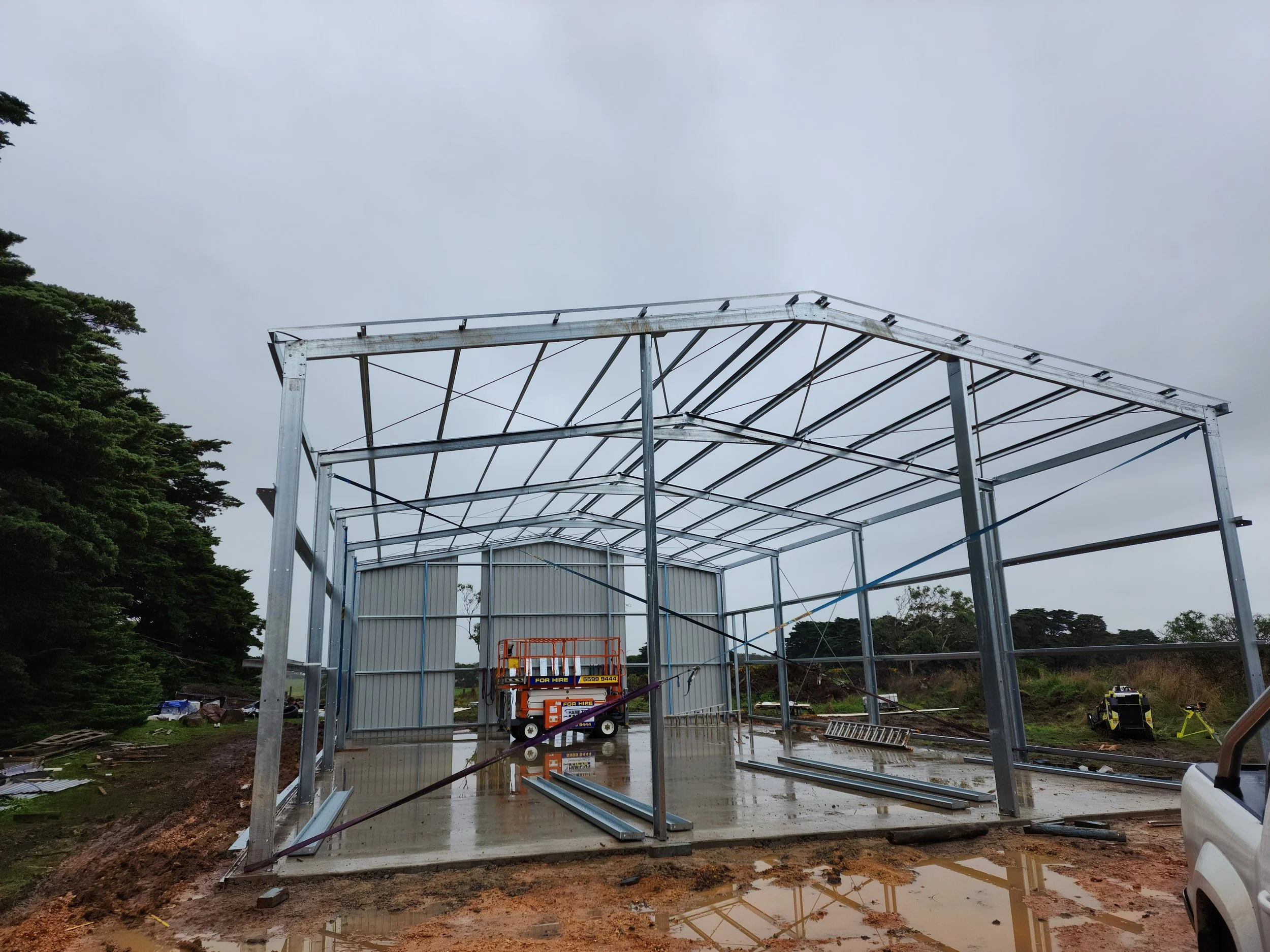 Steel frame structure of a building under construction on a rainy day, with tools and equipment nearby.