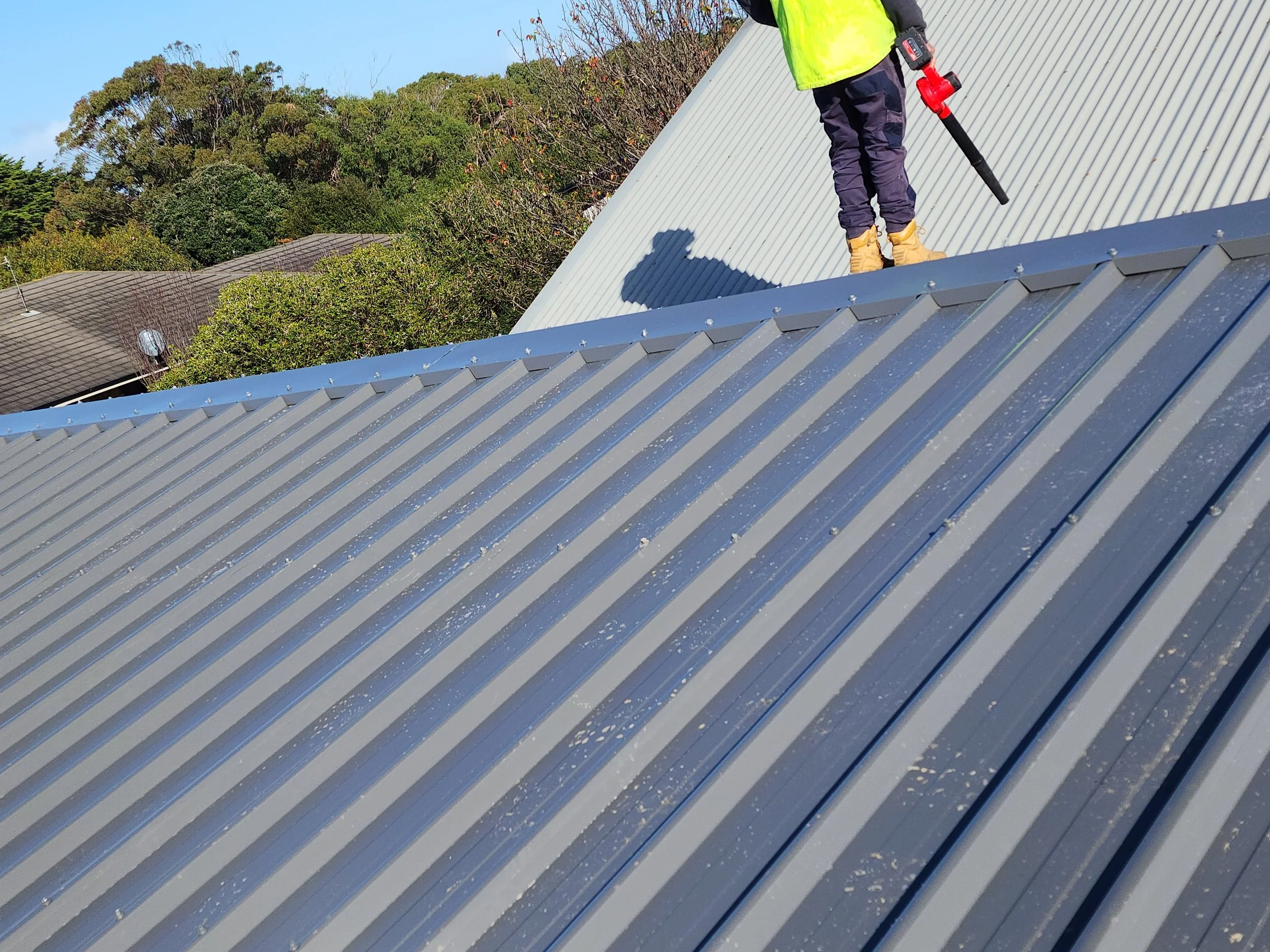 A builder standing on a metal roof with a leaf blower, wearing a bright yellow safety vest, dark pants, and tan boots, with trees and rooftops in the background.