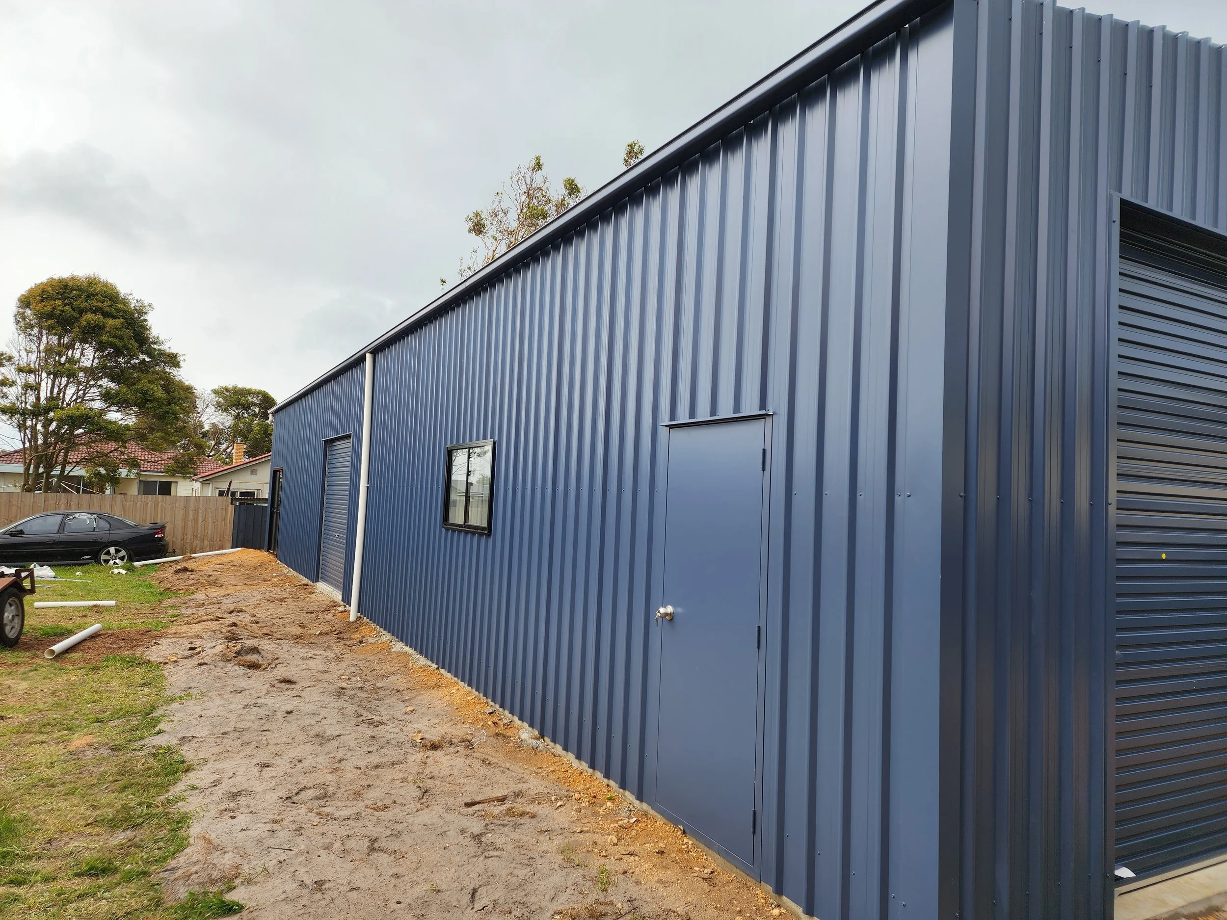 Side view of a blue metal building with a roller door, window, and side door, situated on a partly grassy and dirt lot with a black car in the background and trees behind a wooden fence.